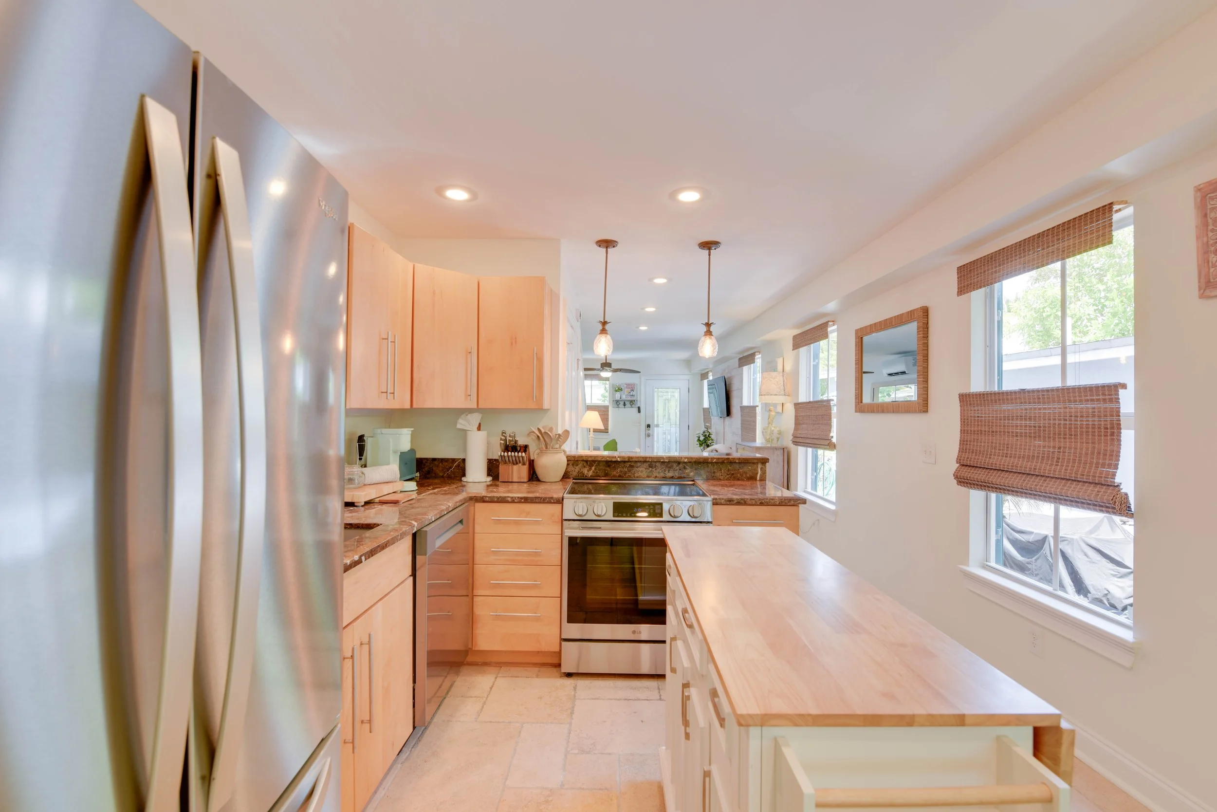 A modern kitchen with light wooden cabinets, a granite countertop, and large windows with woven blinds. Recessed ceiling lights and hanging pendant lights are visible, along with a stainless steel refrigerator and a stove.