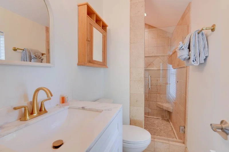 Bathroom with gold fixtures, a white vanity with a marble countertop, a mirror, a wooden wall-mounted cabinet, a toilet, and a glass-enclosed shower with beige tiles.