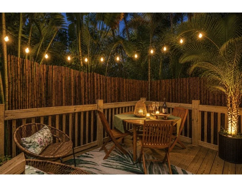 Outdoor wooden deck decorated with string lights, a small round dining table with chairs, a wicker chair with a floral cushion, a potted palm, and candles creating a cozy evening atmosphere.