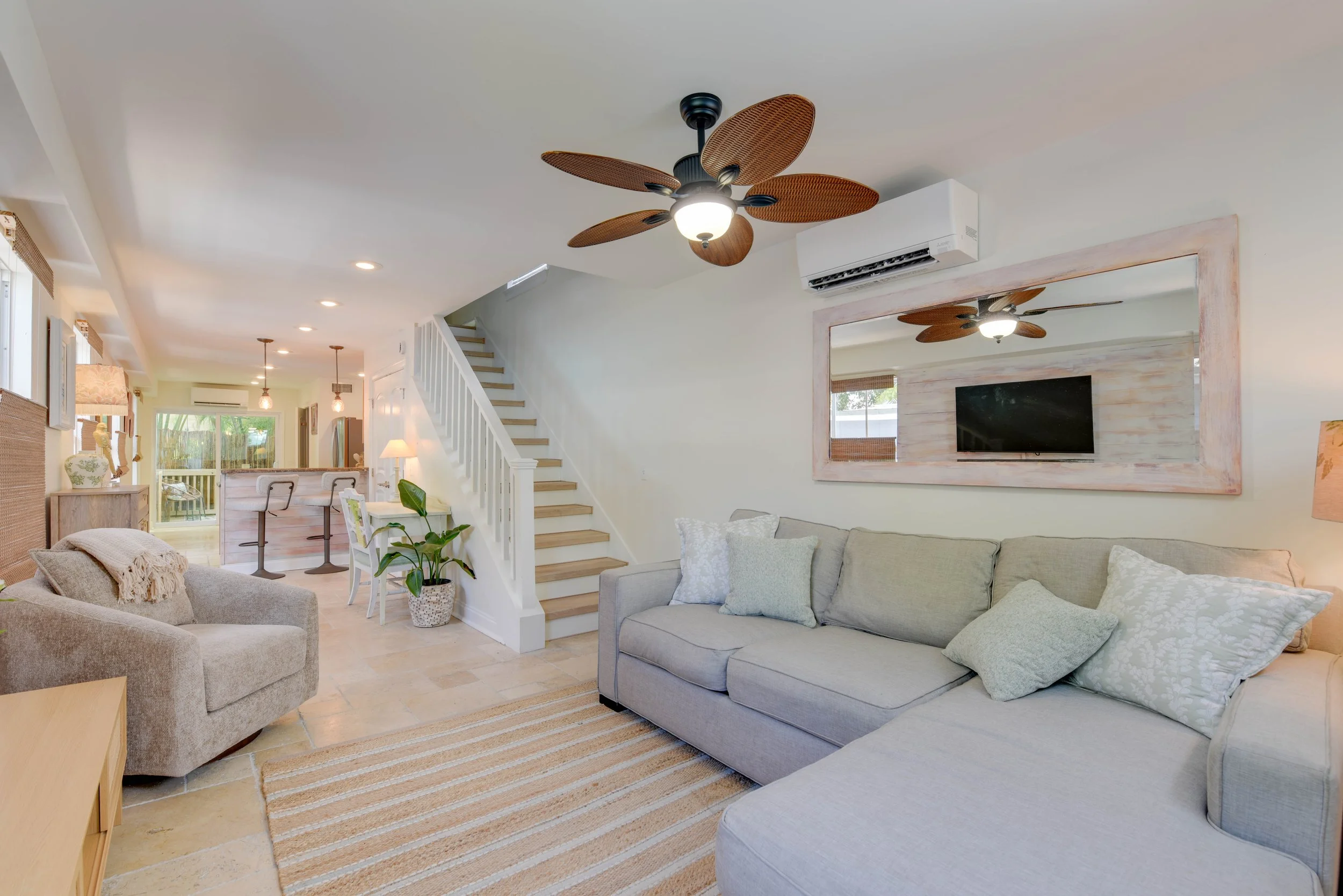 Living room with beige and gray couches, a striped rug, a wall-mounted TV framed in distressed wood, a white staircase, a ceiling fan with leaf-shaped blades, air conditioning unit, windows with bamboo blinds, and a kitchen area in the background.