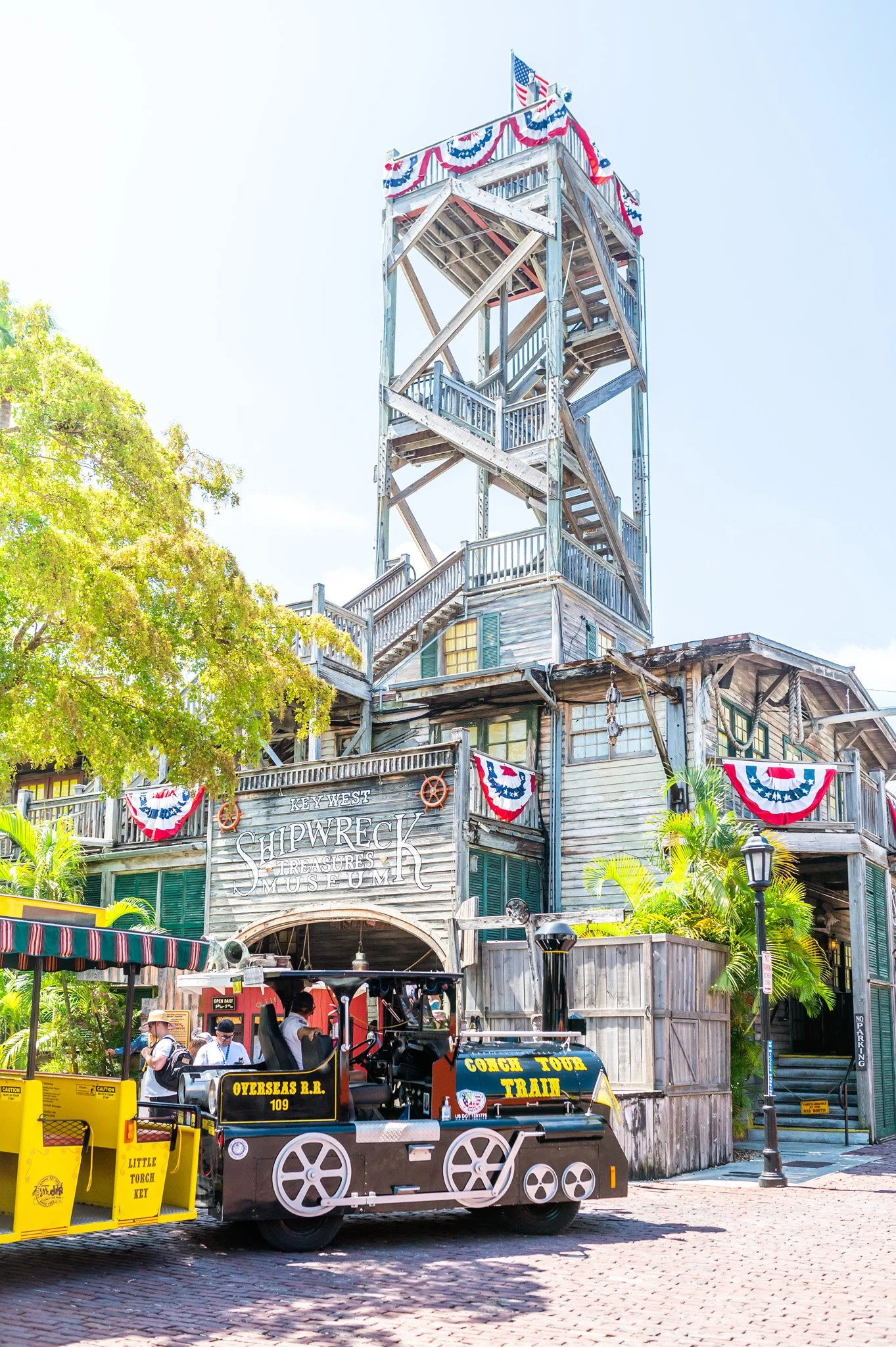 A vintage wooden building with a tower and American flags decorated with red, white, and blue bunting. There is a sign that reads 'Key West Shipwreck & Treasures Museum' and a tour train in the foreground labeled 'Cocoa Tour Train' and 'Overseas R.R. 109'. People are walking near the train and the building.