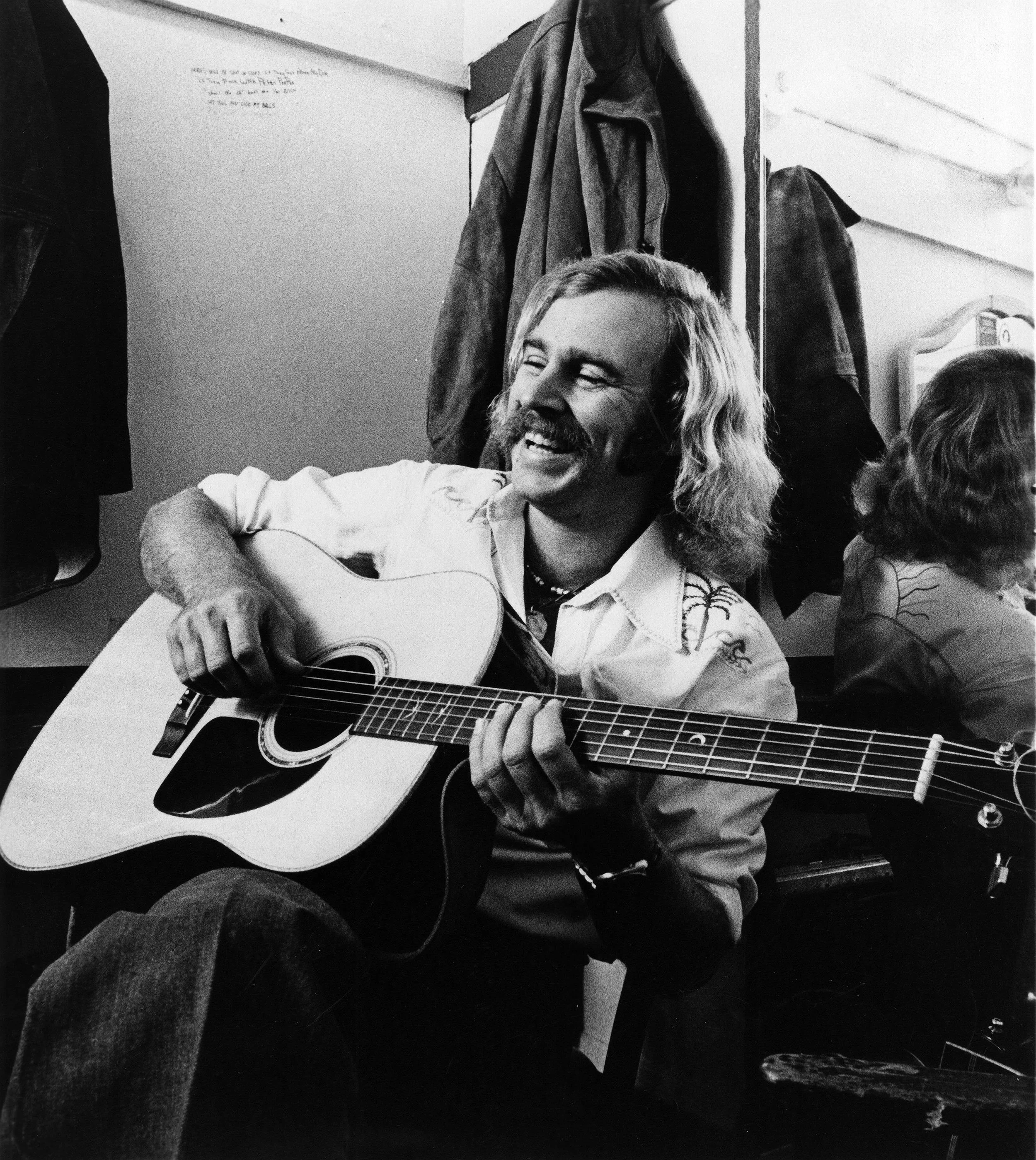 Black and white photograph of a man with long hair and a beard playing an acoustic guitar, smiling, in an indoor setting with clothing items hanging on the wall behind him.