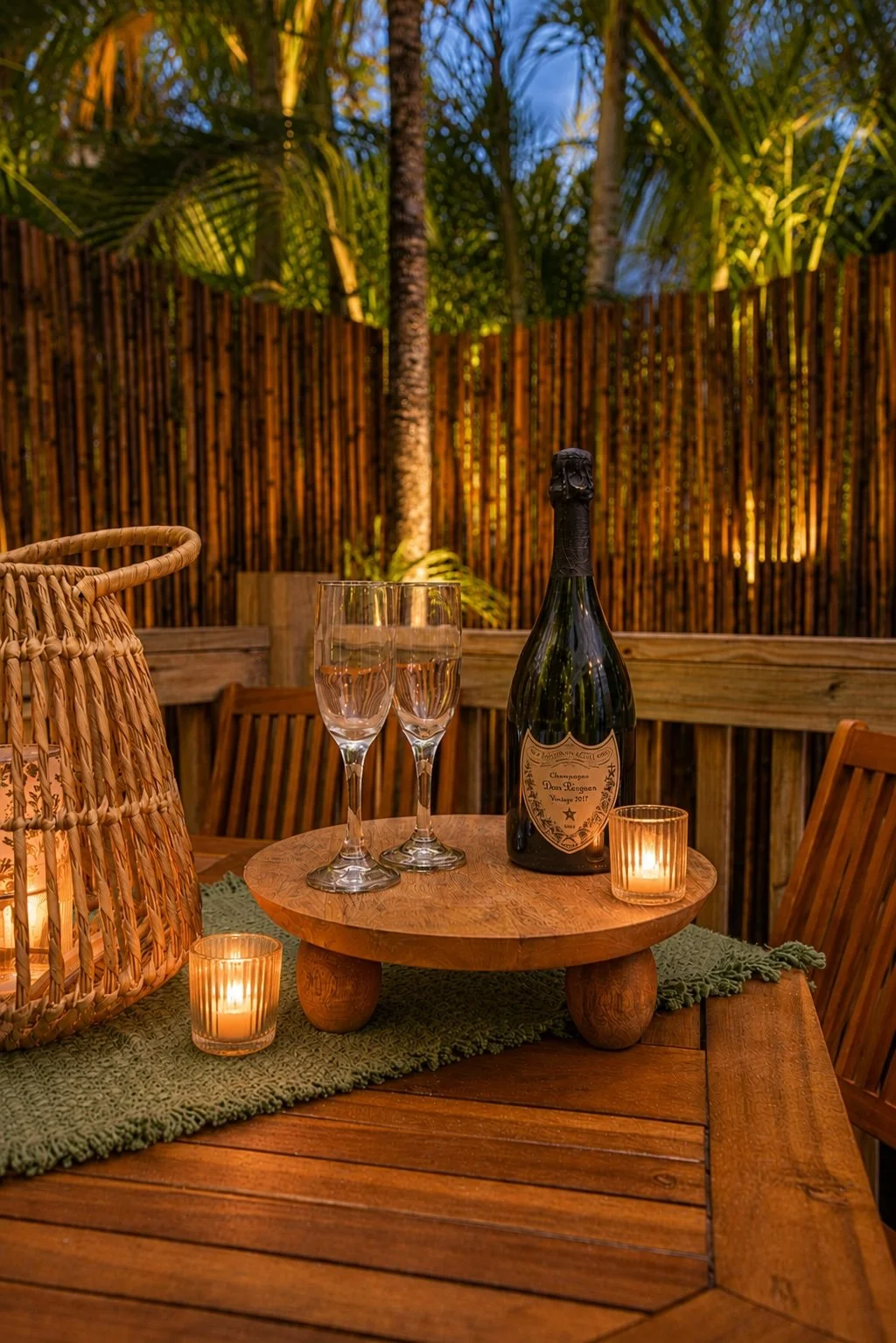 A wooden outdoor table decorated for celebration with two champagne glasses, a bottle of champagne, a candle, and a wicker basket, with a tropical garden and bamboo fence in the background.