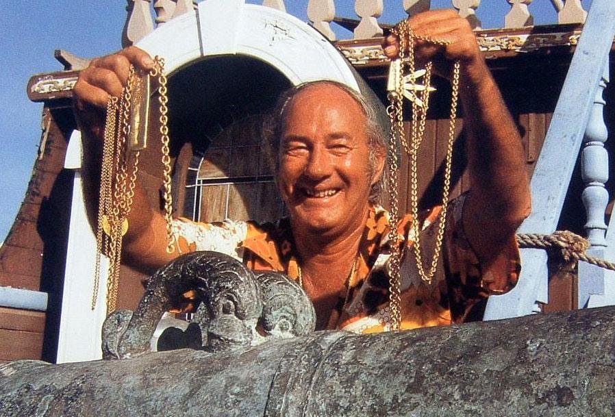 Smiling man holding gold chains, standing behind a stone railing with a sculpted pony underneath, in front of a colorful wooden structure with a staircase.