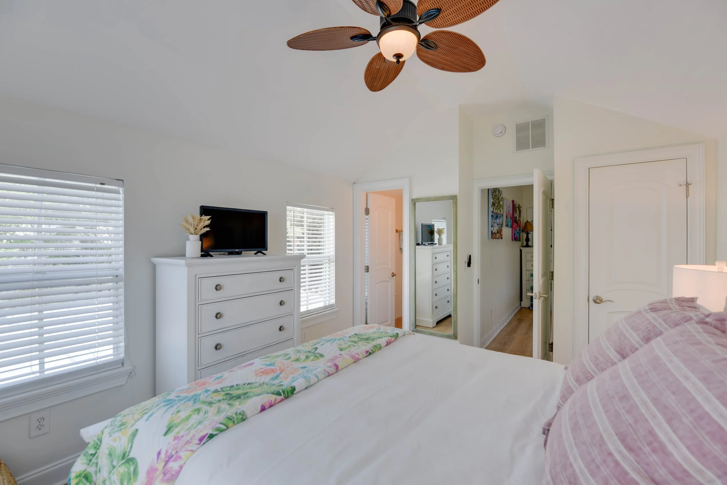 A bedroom with a bed covered in a white duvet and a colorful floral blanket, pink and white striped pillows, a white dresser with a small TV and a vase with dried flowers, large windows with white blinds, a standing mirror, and an open door leading t