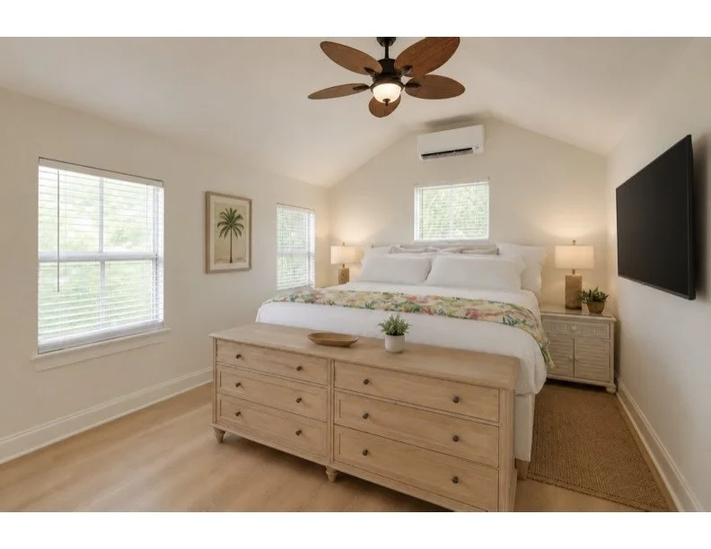Bright bedroom with a large bed, wooden dresser at foot, nightstands with lamps, window with blinds, wall art, ceiling fan, air conditioning unit, and wall-mounted TV.