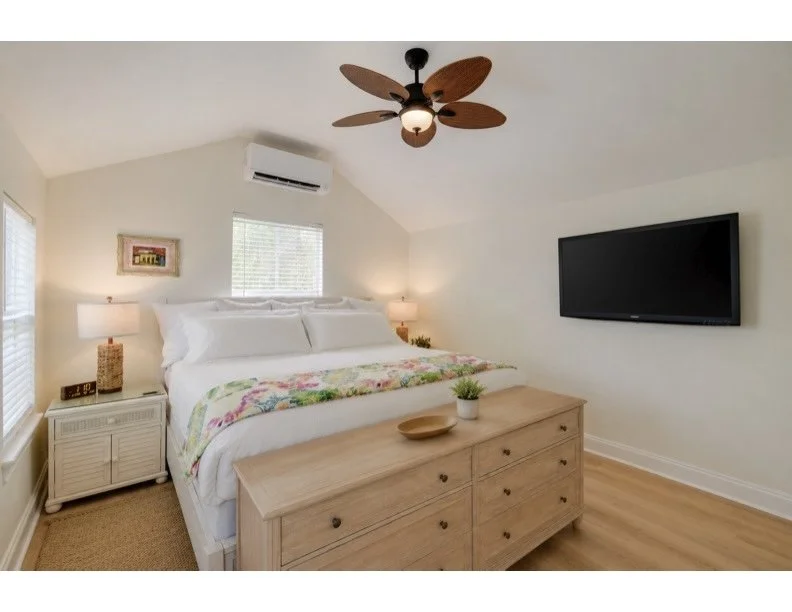 A bedroom with a white bed, colorful floral blanket, wooden dresser, wall-mounted flat-screen TV, ceiling fan with wooden blades, two nightstands with lamps, and windows with blinds.