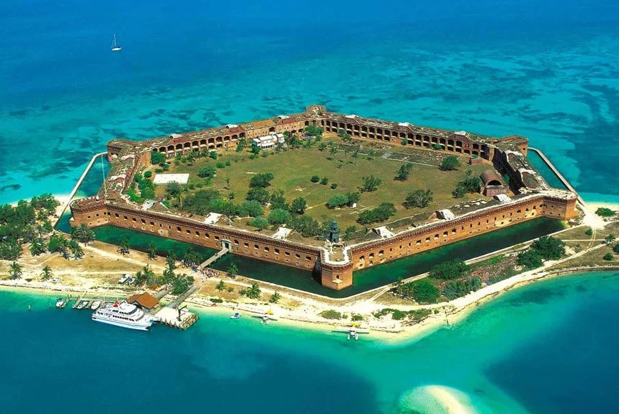 Aerial view of a large historic fort on a small island surrounded by turquoise ocean, with a docked boat and lush greenery inside the fort