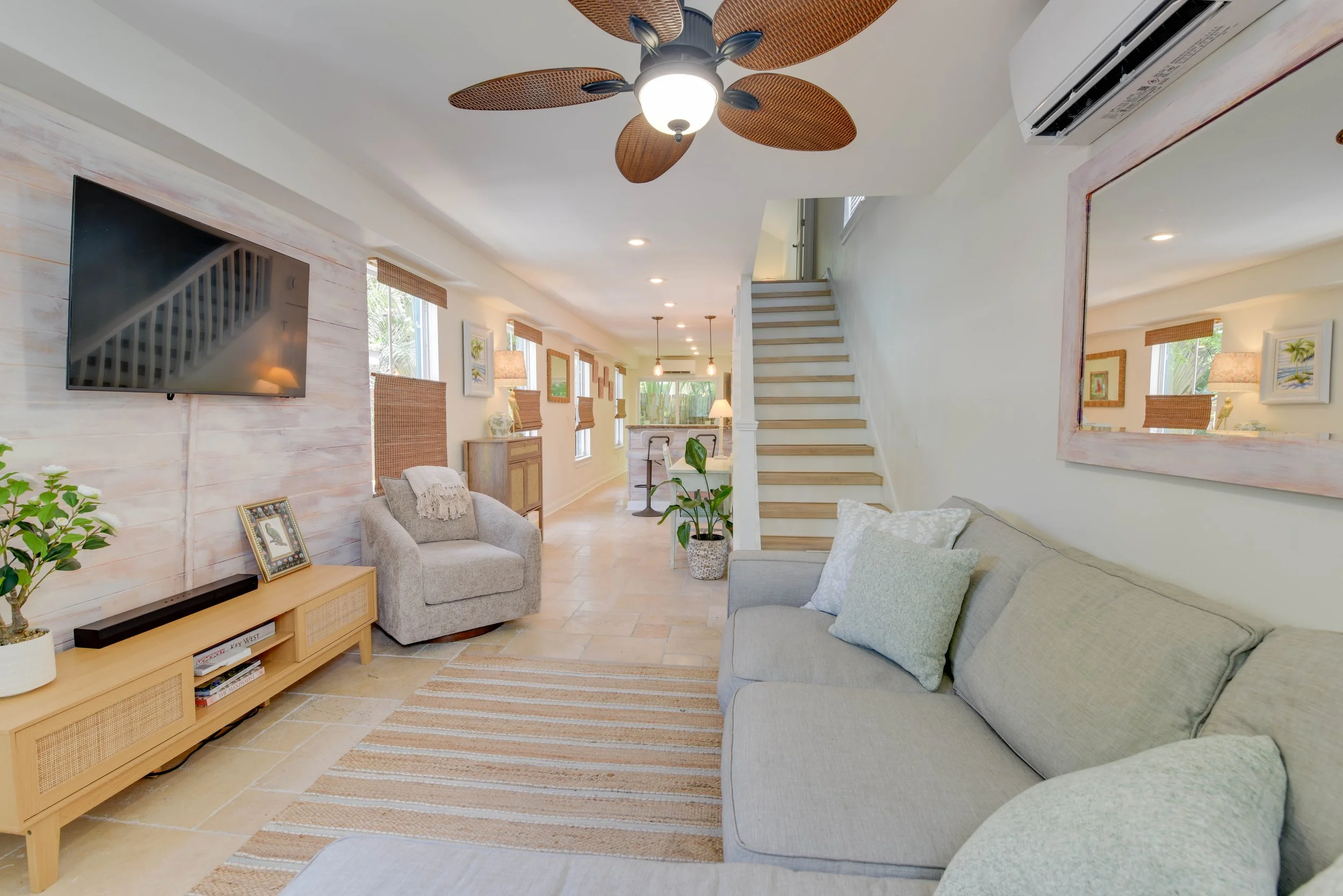 Living room with beige sofas, a wooden TV stand, framed pictures, a ceiling fan with leaf-shaped blades, and an air conditioning unit on the wall.