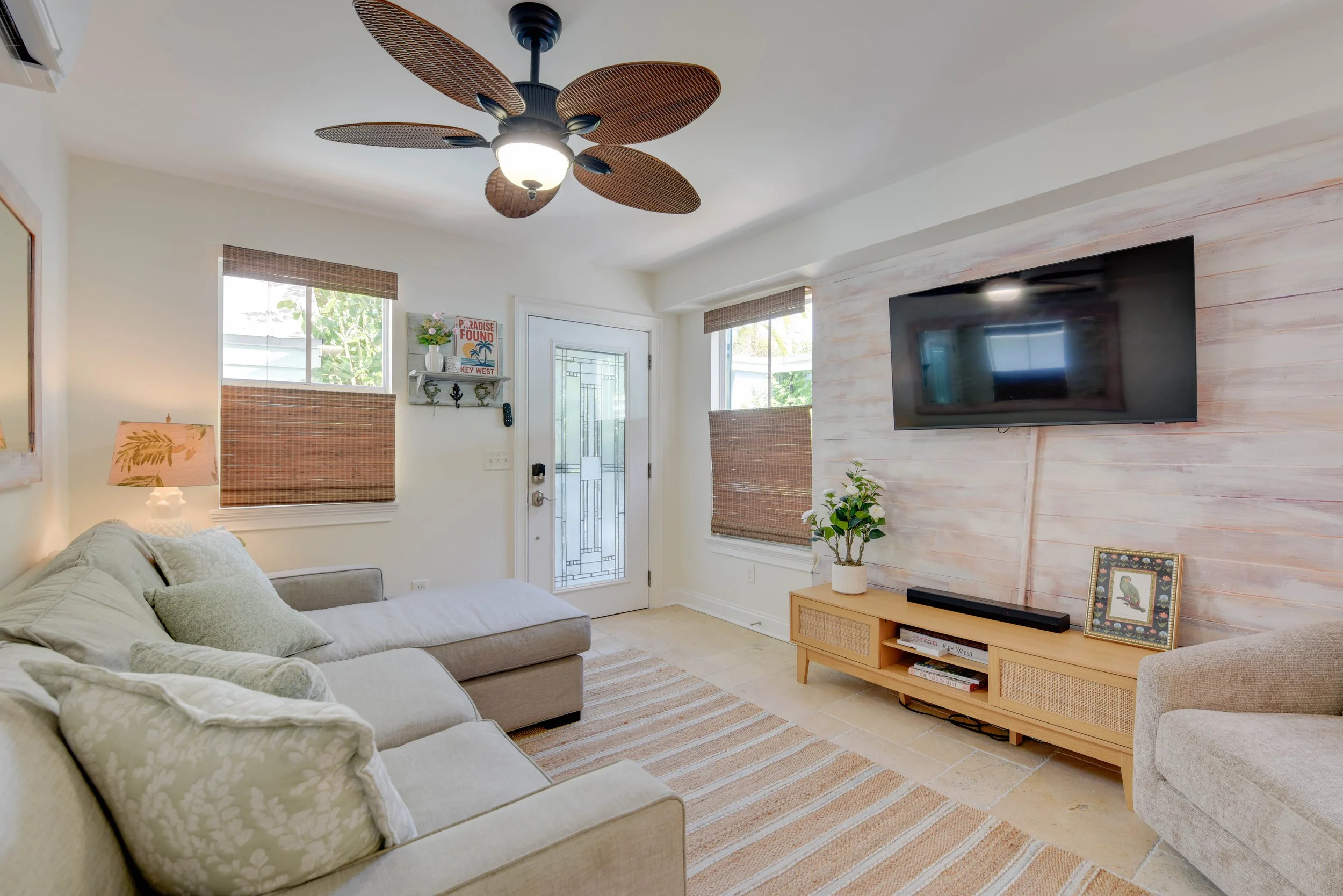 Living room with a gray sofa, beige armchair, wooden TV stand, wall-mounted TV, wood-paneled wall, ceiling fan with leaf-shaped blades, two windows with woven blinds, and various decorative items including a framed picture and potted plants.