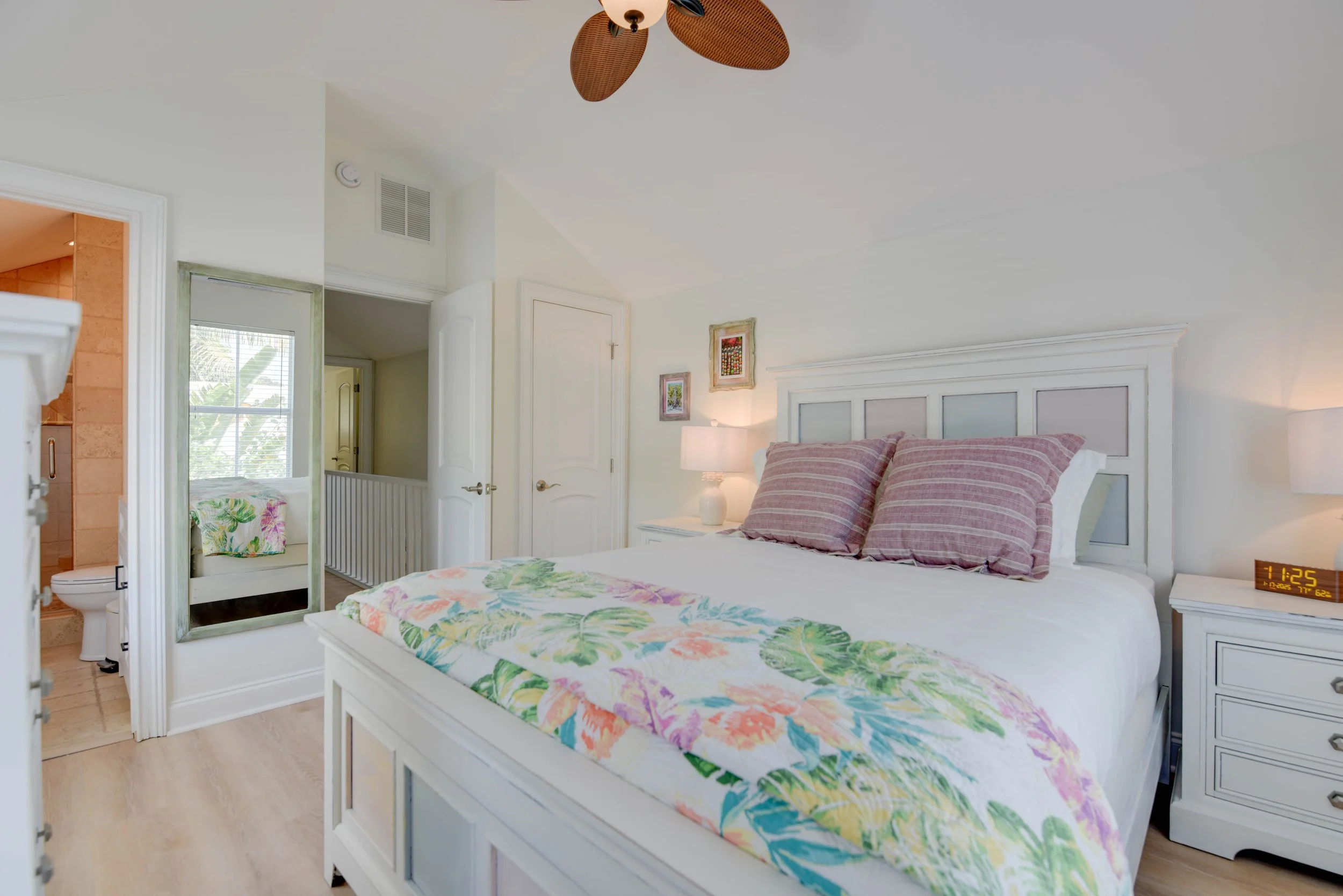 A bedroom with a white wooden bed frame featuring floral bedspread and matching pillows. There is a nightstand with a lamp and digital clock. The ceiling has a ceiling fan with wooden blades. An open door reveals an adjacent bathroom with a visible t