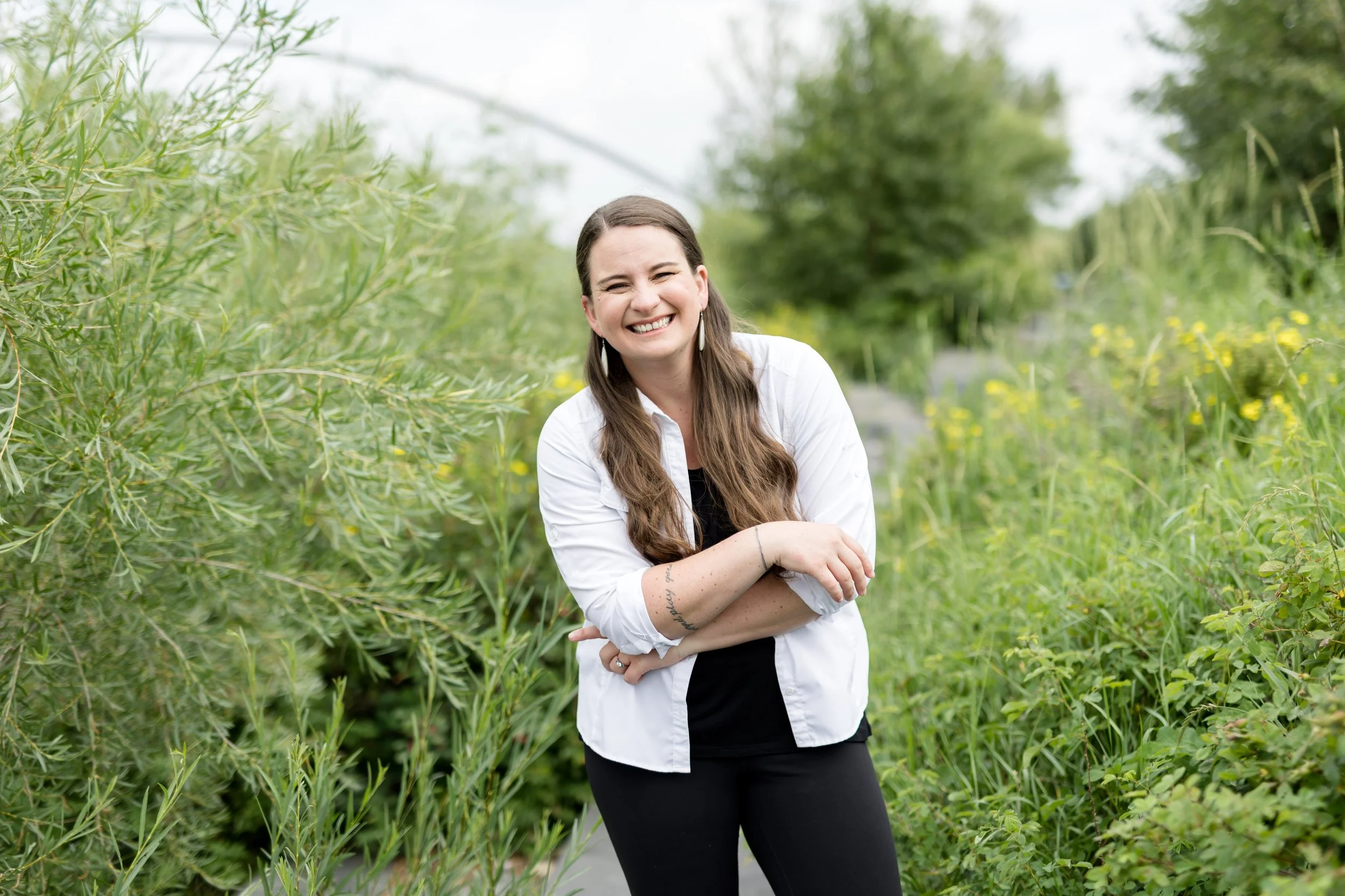 A woman with long brown hair, wearing a white shirt and black pants, smiling with her arms crossed, standing among green plants and trees.