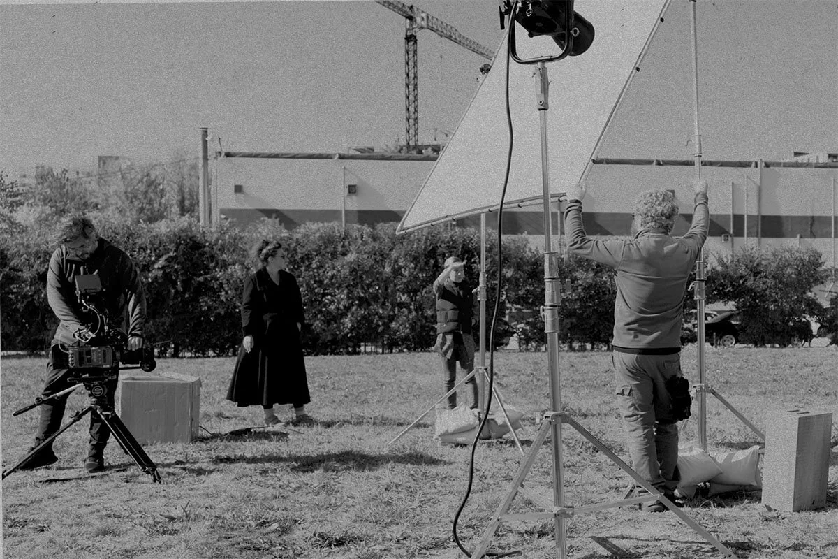 Black and white photo of a film crew setting up a scene outdoors with three actors, one behind a camera, one adjusting lighting, and the others standing, with trees and a building in the background.