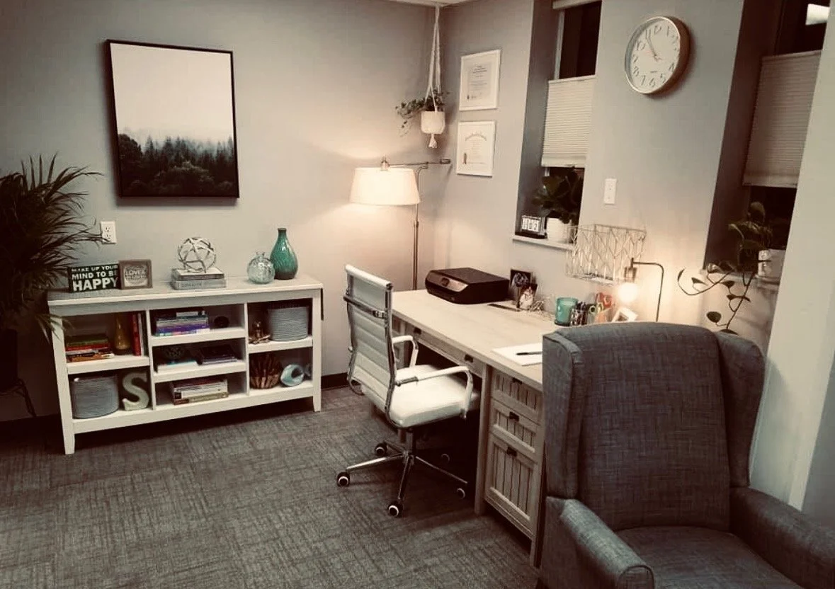 Office space with desk, white office chair, gray armchair, bookshelf, decorations, and wall clock.