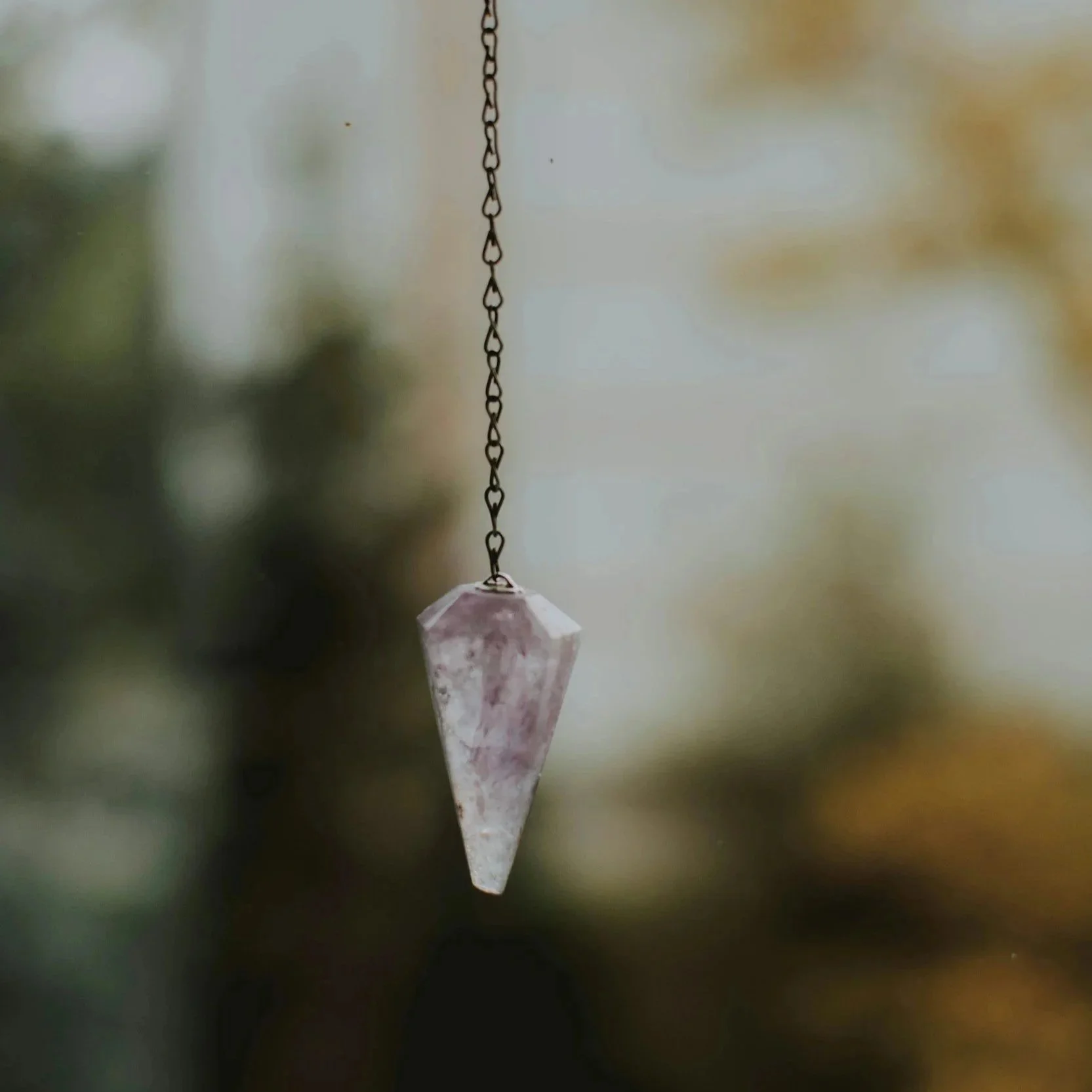A close-up of a crystal pendulum hanging from a chain with a blurred background.