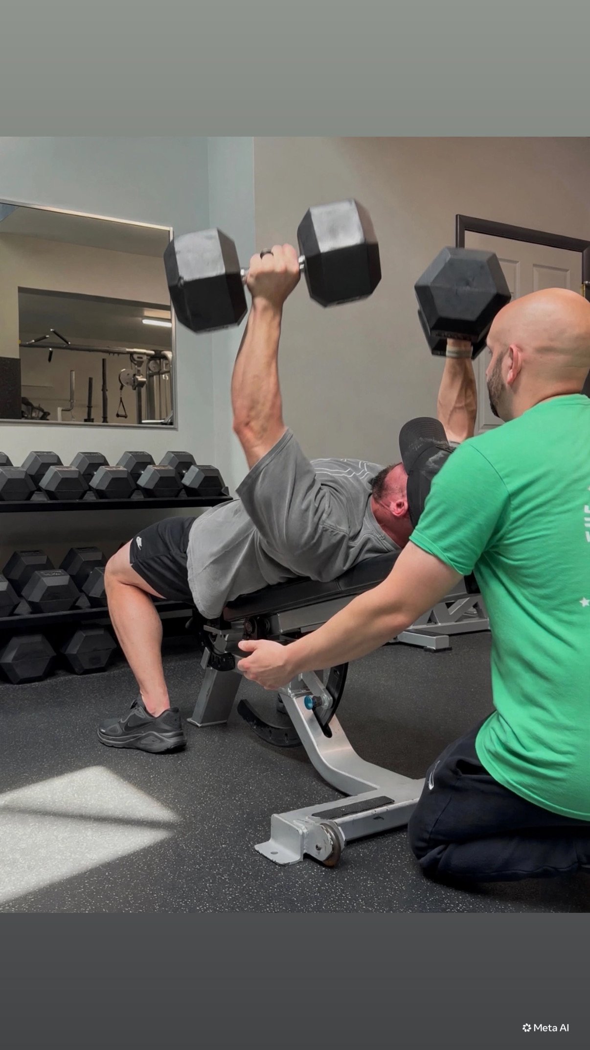 A man lying on a workout bench lifting two black dumbbells, with a trainer assisting him, in a gym with weight racks and mirrors.