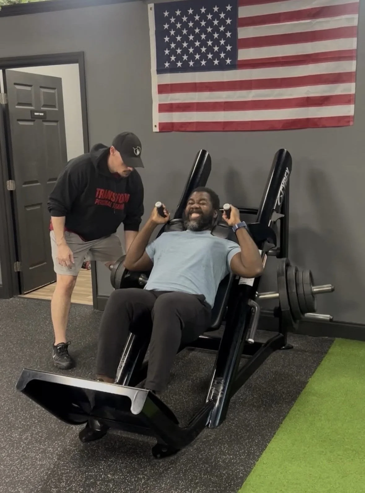 A man sitting on a leg press machine in a gym, smiling as he lifts weights. Another man stands beside him, leaning over and smiling. An American flag is hanging on the wall behind them.