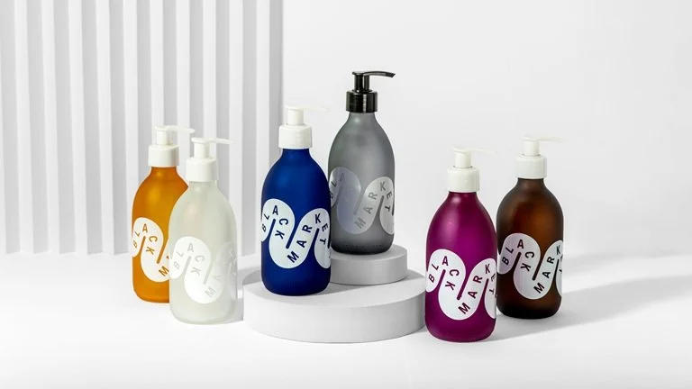 Six colorful bottles of skincare or cosmetic products displayed on a white surface with a gray and white background, one with a pump dispenser and the others with screw caps.