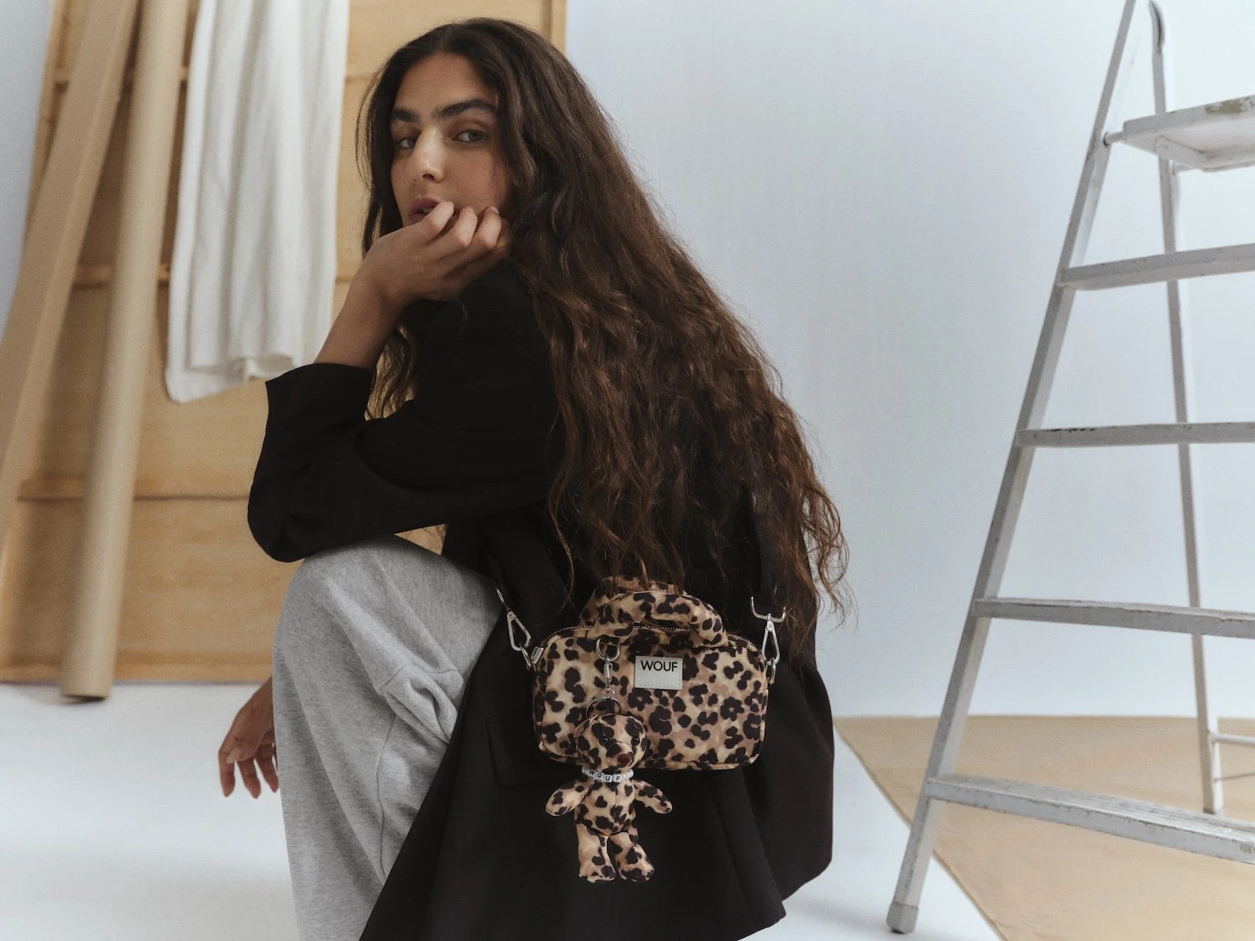A young woman with long, curly dark hair looking at the camera, sitting indoors. She is wearing a black top and gray sweatpants. She has a small leopard-print backpack hanging from her arm.