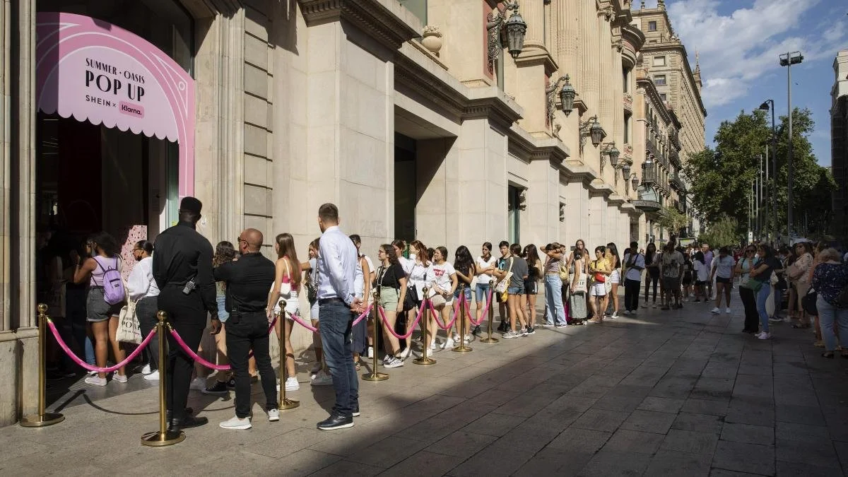 People standing in line outside a store with a pink sign that reads 'Summer - Oasis Pop Up' in front of a large, ornate building on a city street.