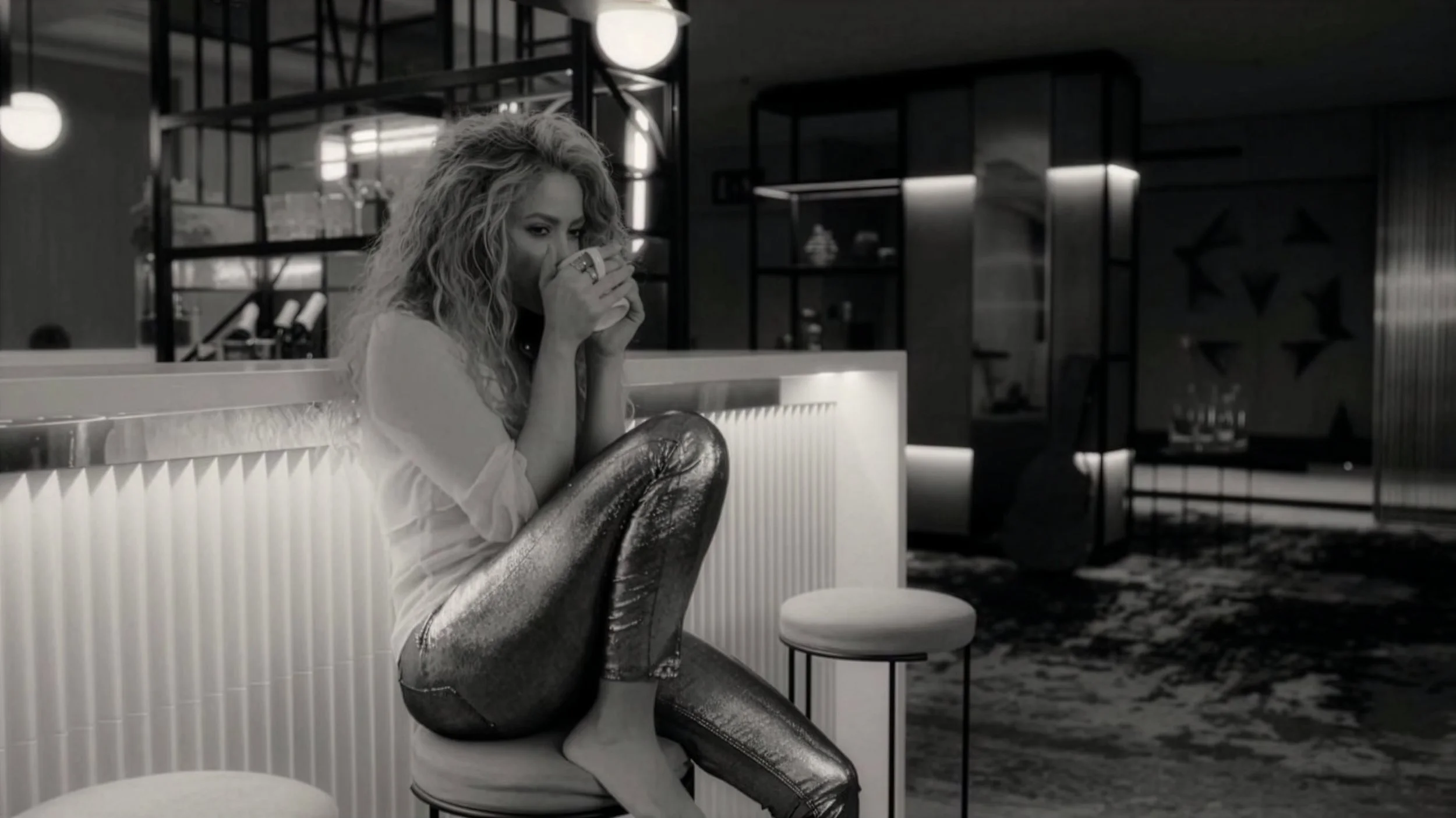 A woman with curly hair sitting in a modern, stylish room, holding a cup, with a relaxed pose and partial smile, in black and white.