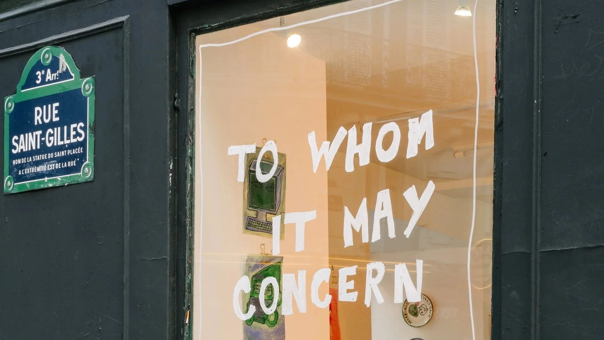 Storefront window with painted text reading "To Whom It May Concern" and a small drawing of a green computer. A street sign indicates the location as Rue Saint-Gilles in the 3rd arrondissement.