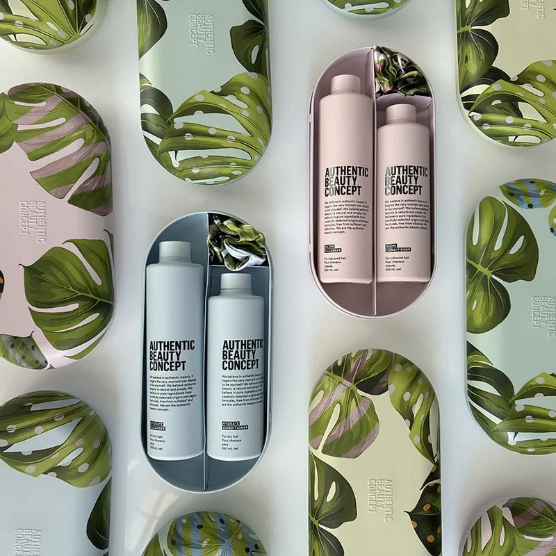 Flat lay of skincare products labeled 'Authentic Beauty Concept' in sleek bottles, set inside eco-friendly, botanical-themed packaging with green monstera leaves on a white background.