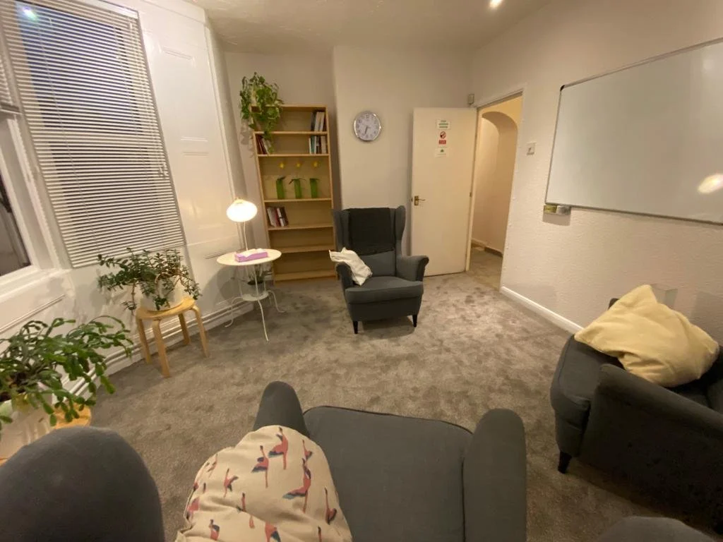 A cozy living room with gray armchairs, indoor plants, a white side table with a lamp, a gray carpet, a bookshelf, a whiteboard on the wall, and a doorway leading to another room.