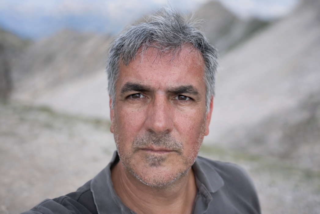 Close-up of a middle-aged man with gray hair and a gray beard, standing outdoors in a natural landscape with hills and a cloudy sky.