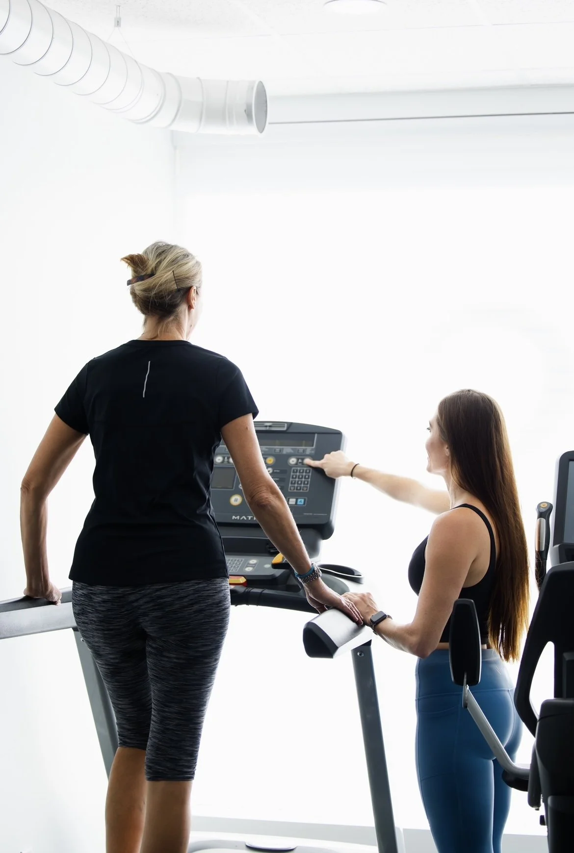 Two women working out on treadmill and elliptical in a brightly lit, modern gym.