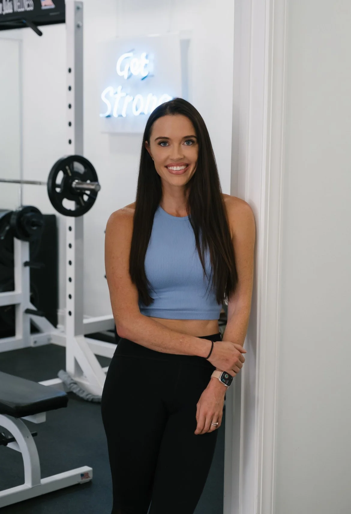A woman standing in a gym, wearing a blue workout top and black leggings, smiling at the camera with a neon sign that says 'Get Strong' in the background.