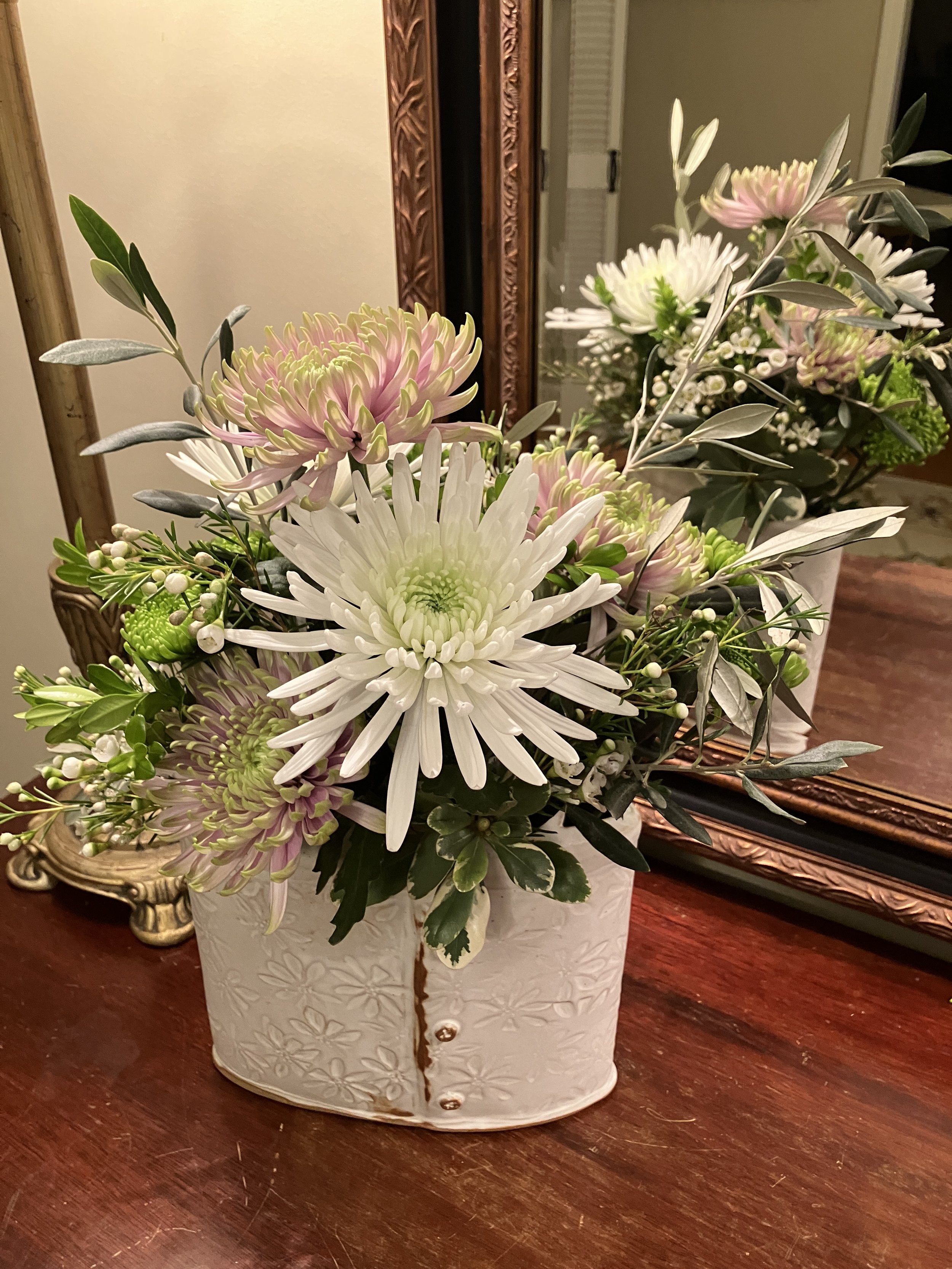 A Margaret Barber white ceramic vase with embossed floral pattern containing a bouquet of white and pink flowers with green leaves, placed on a dark wooden table. A large framed mirror with ornate frame is visible in the background.