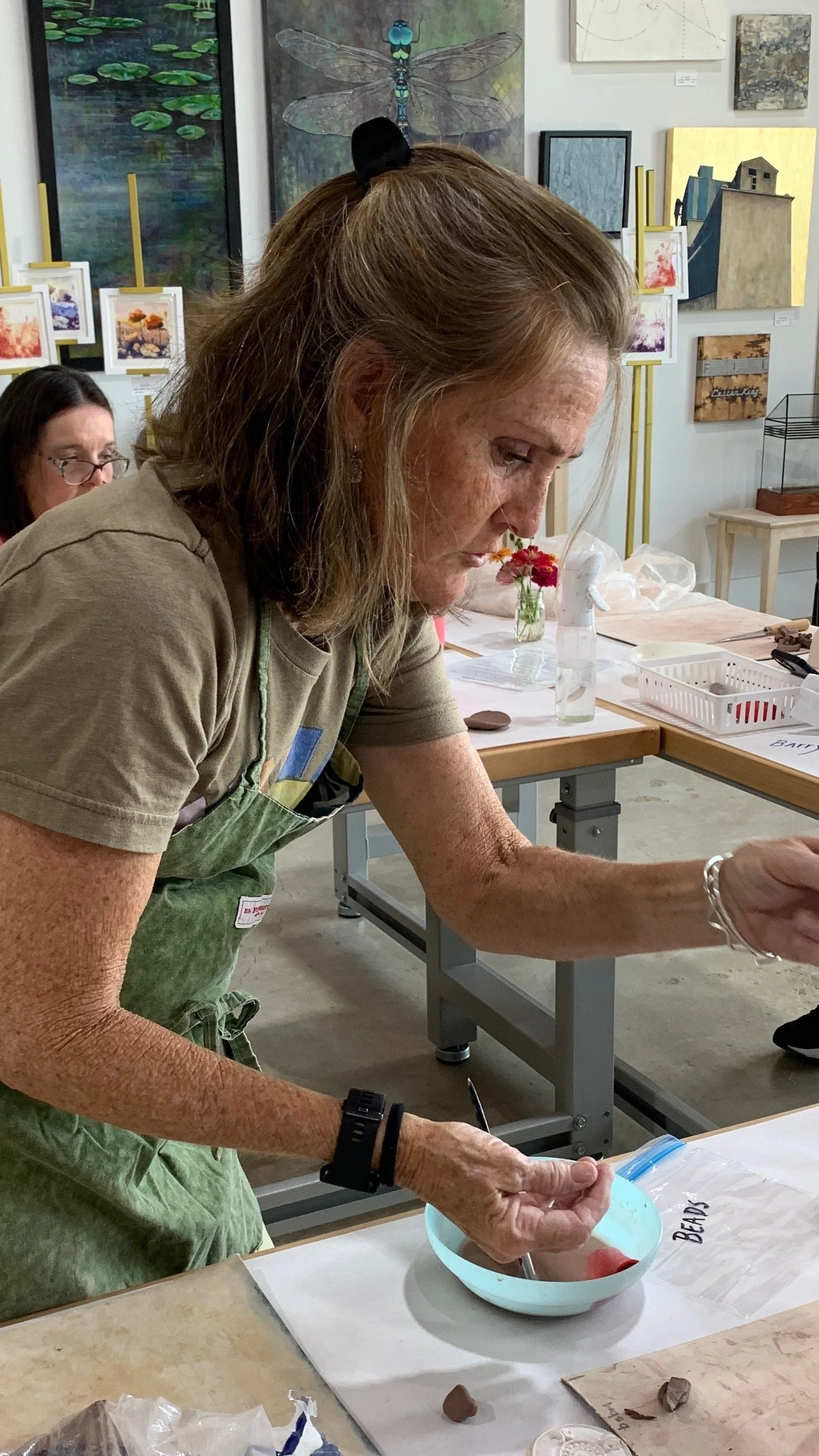 A woman with red hair and freckles, wearing a green apron, is working with clay at a table at 3 Houses Studio, Gluckstadt, MS. Behind her, there are paintings on the wall, including a large dragonfly and various abstract and landscape artworks.