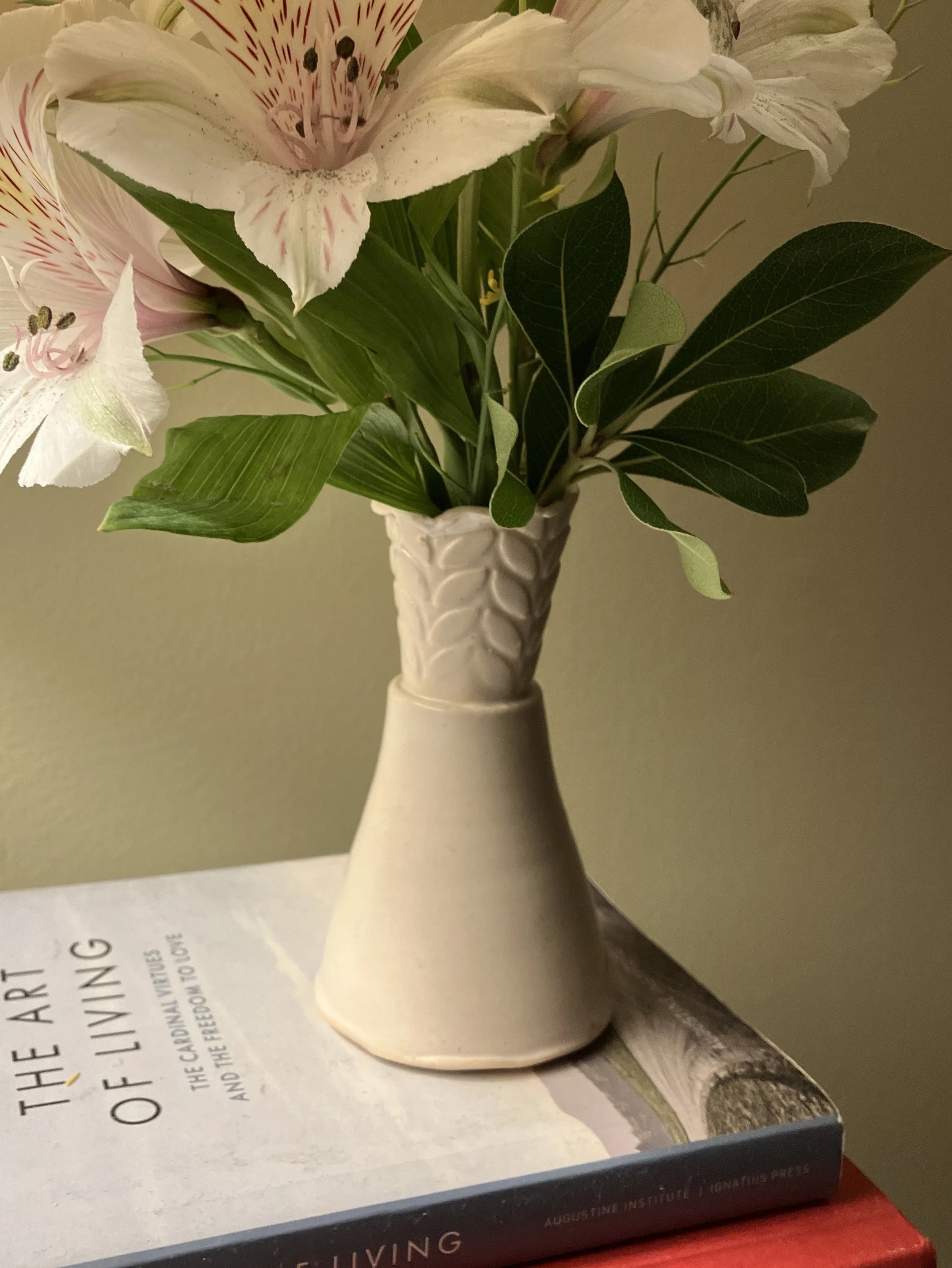 A Margaret Barber ceramic vase with a textured pattern containing a bouquet of white lilies with pink and purple streaks and dark stamens, placed on a table with a book underneath.
