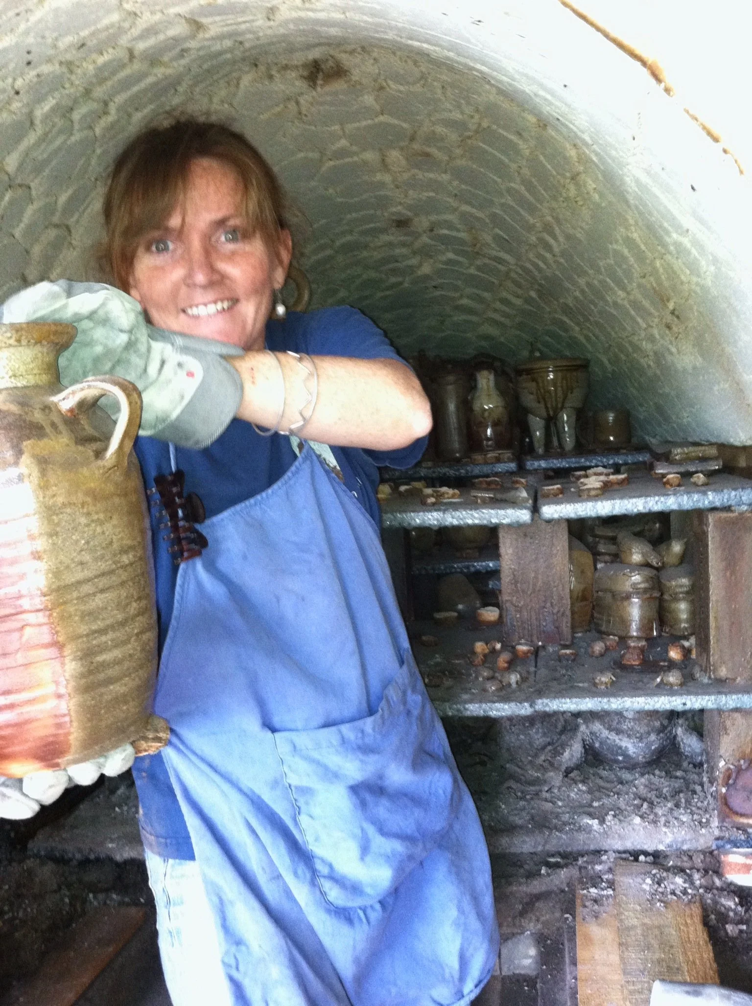 A woman wearing a blue apron and gloves inside a Penland kiln, smiling and unloading pottery items from kiln in the background.