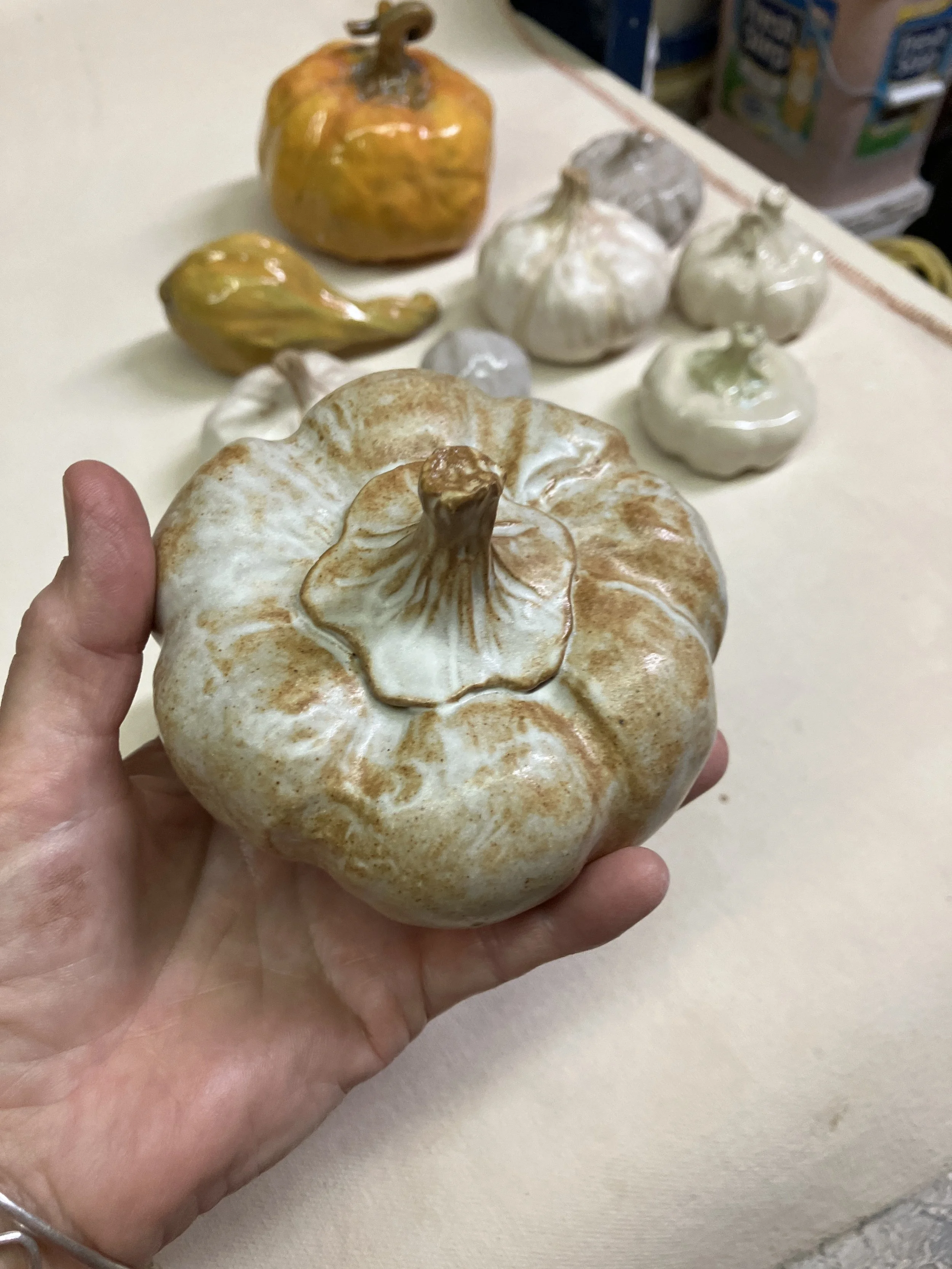 A person holding a Margaret Barber decorative pumpkin with a brown stem, surrounded by other ceramic gourds on a canvas surface.