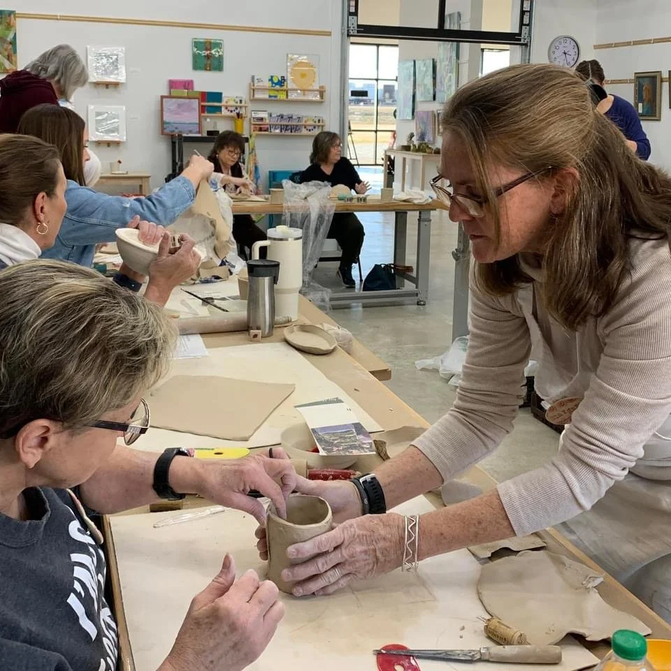 People participating in a Margaret Barber pottery class, Margaret helping a woman shape a clay pot in a bright studio filled with artwork and art supplies.