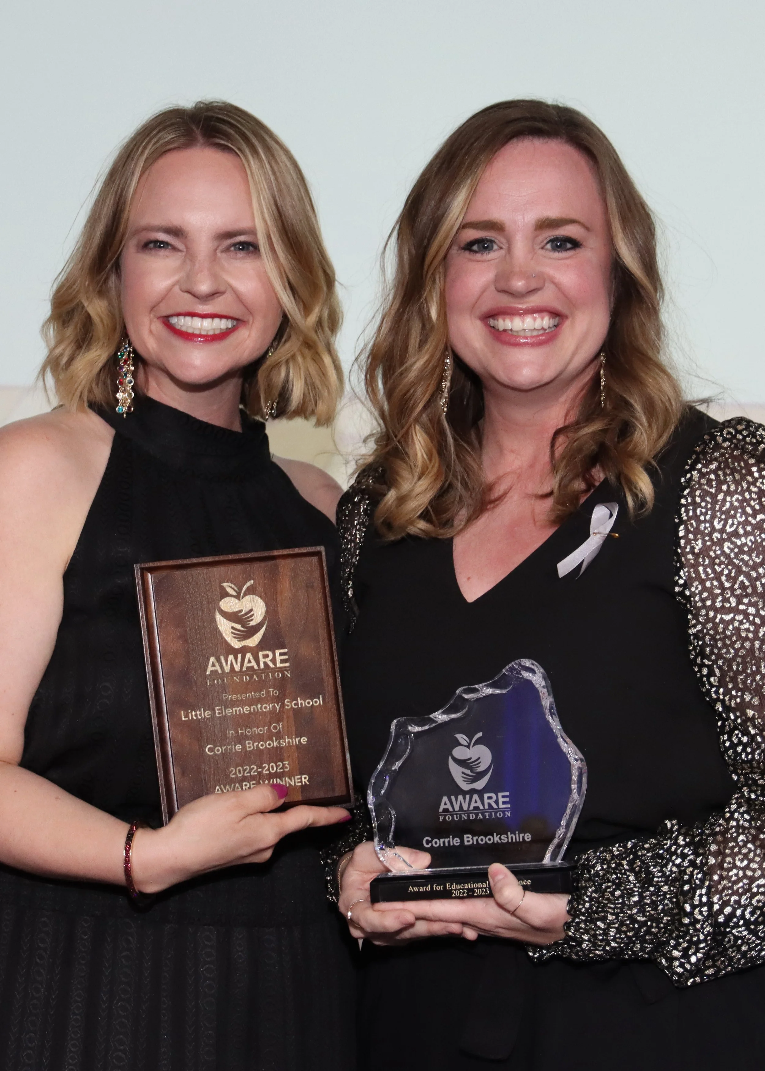 Two women are smiling and holding Aware awards at a ceremony. One wears a black sleeveless dress; the other wears a black top with silver sequined sleeves. The woman on the left holds a wooden plaque; the woman on the right holds a glass award.