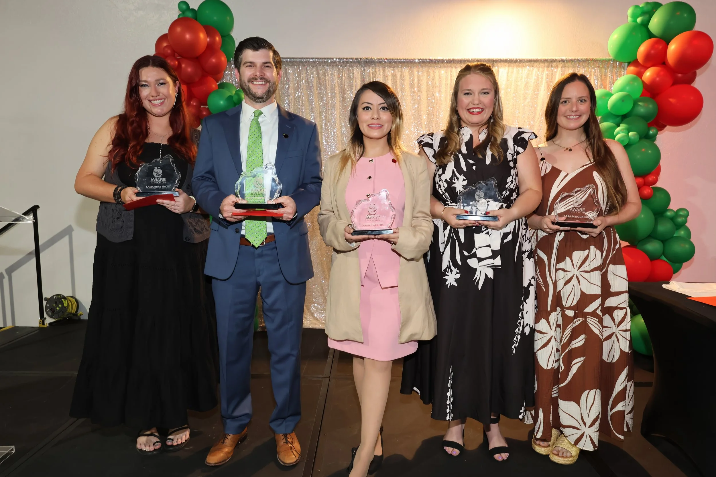 Five people standing in a line, smiling, at the Aware annual awards ceremony with balloons and a shimmering backdrop, each holding an Aware award.