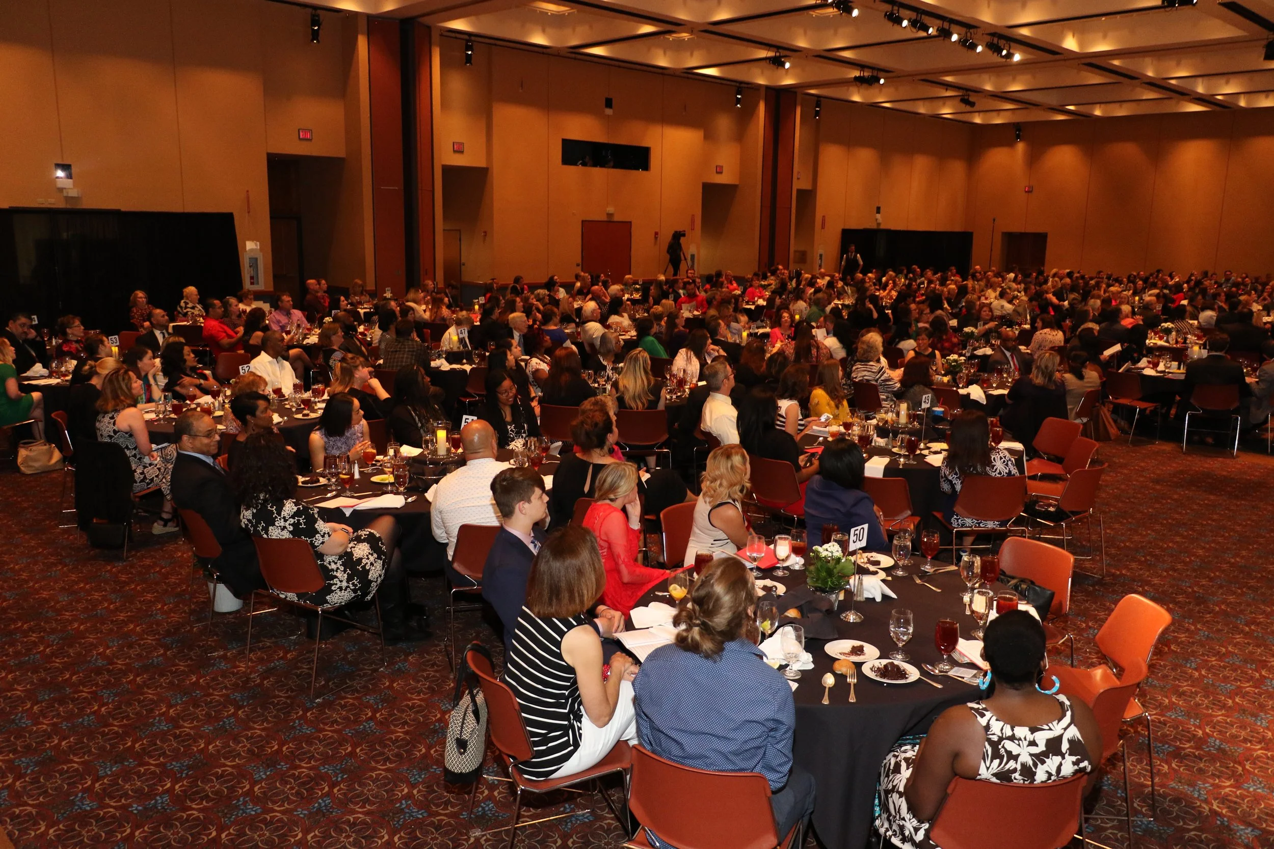 A large banquet hall filled with numerous round tables, each set with tableware, glasses, and small floral centerpieces. The room is crowded with diverse attendees seated and engaged in conversation at the annual Aware awards banquet.