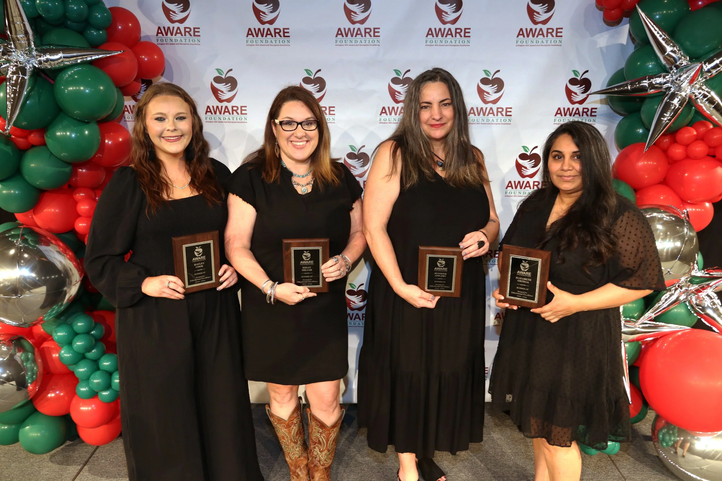Four women standing in front of a backdrop with the AWARE Foundation logo, surrounded by red, green, and silver balloons, holding Aware plaques at an event.