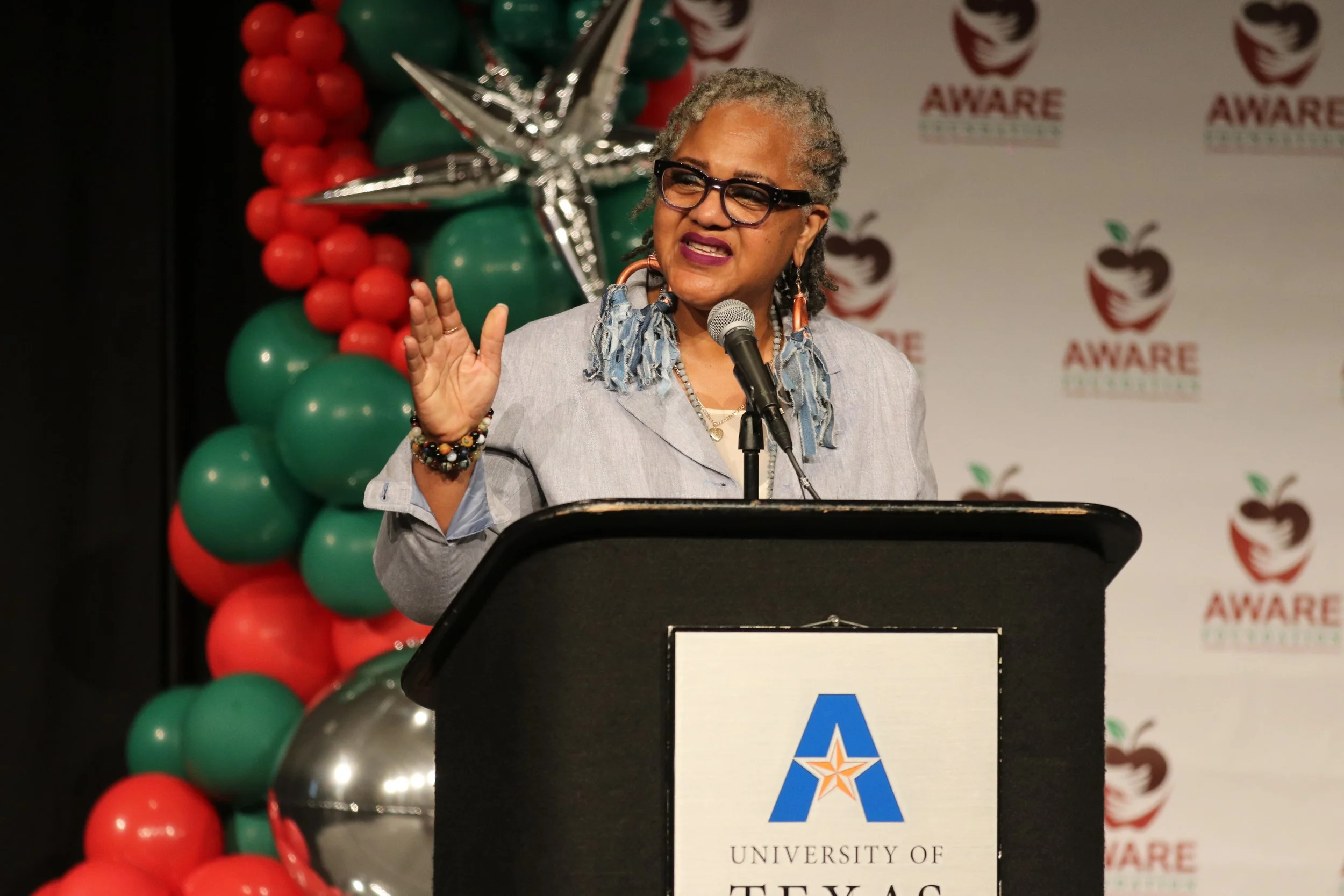 A woman at a podium with a microphone at the annual Aware awards event. Behind her is a decorative balloon arch with red, green, and silver balloons, and a backdrop with a logo and text that reads "AWARE" and an outline of a face in profile.