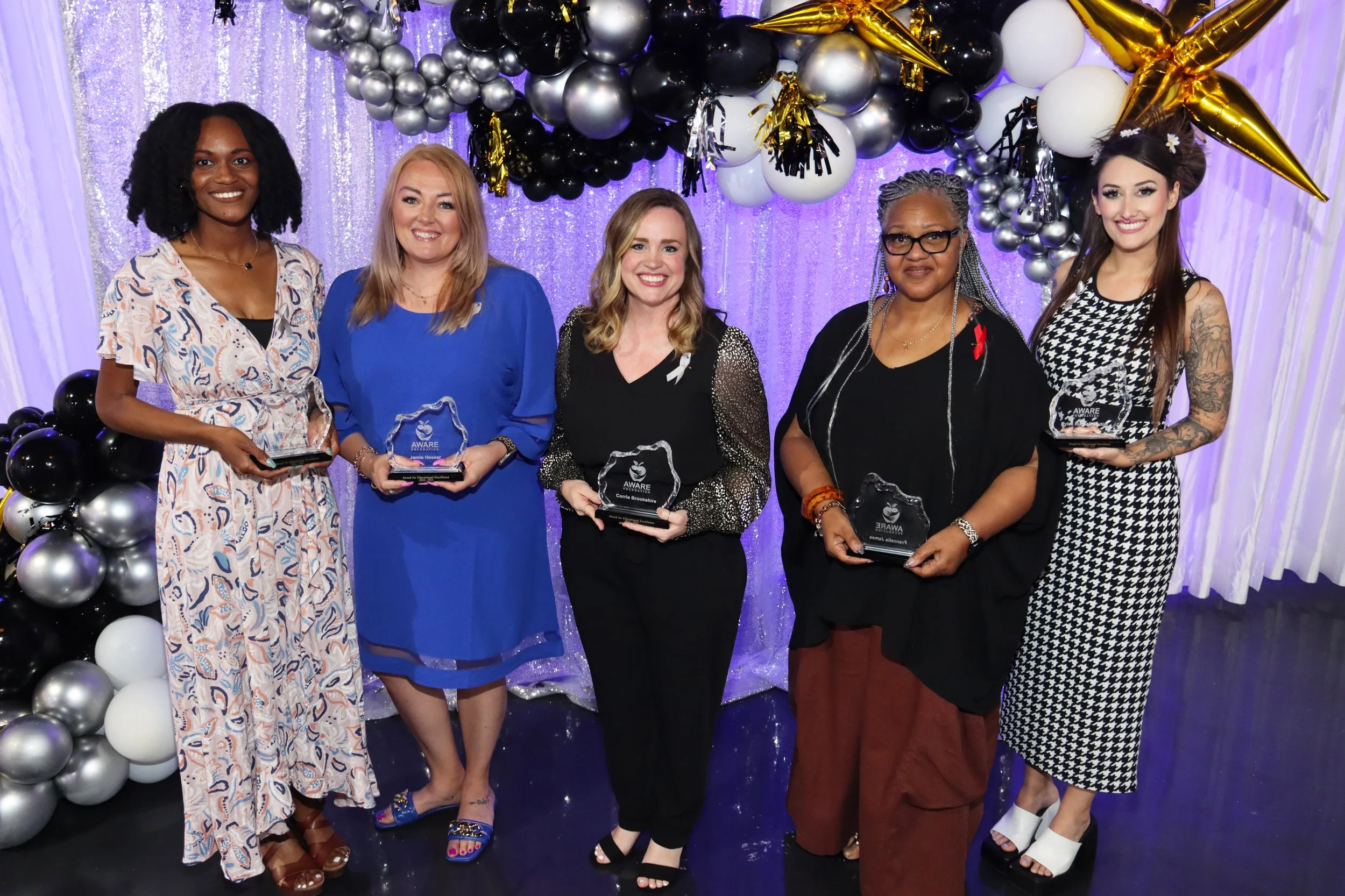 Five women standing in front of a backdrop decorated with black, silver, white, and gold balloons, holding Aware awards, smiling at the camera.
