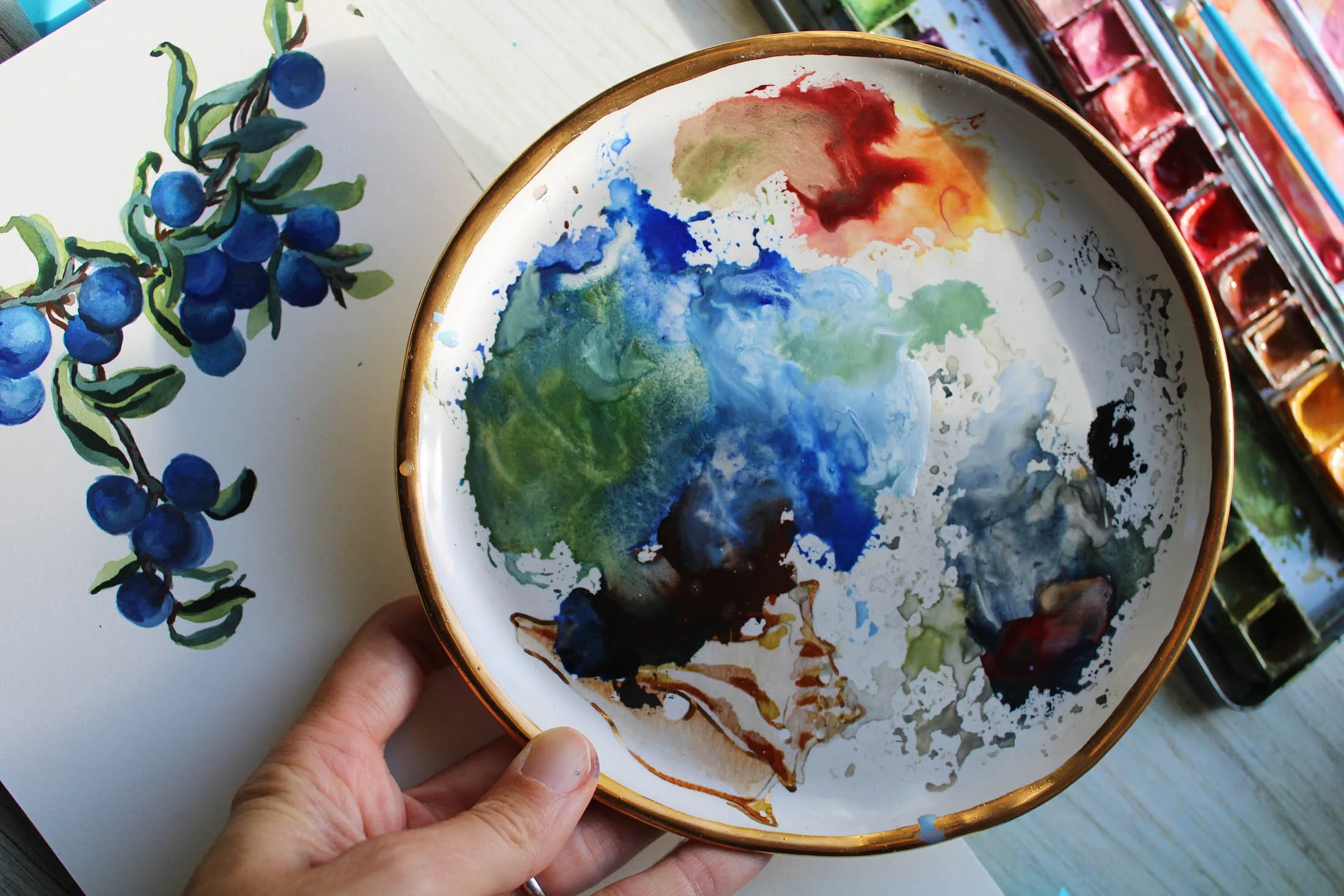 Hand holding a small white ceramic bowl with a gold rim filled with watercolor paint swirls of green, blue, red, black, and brown.
