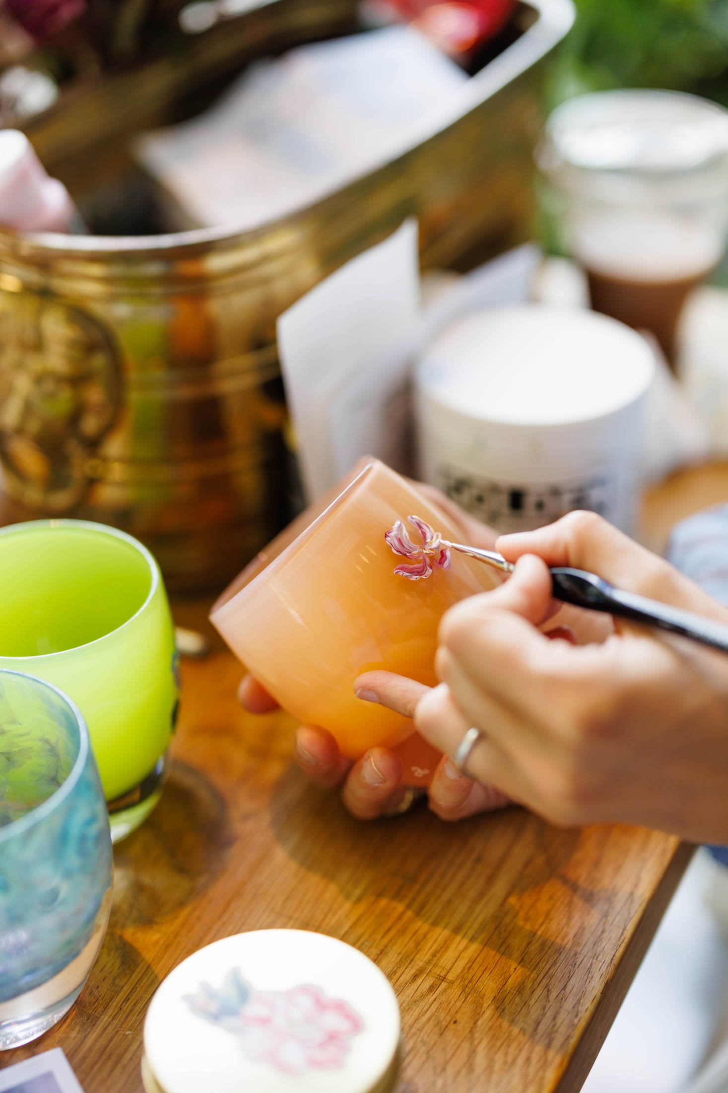 Close-up of a person's hands decorating a pink candle with a flower-shaped ornament. Several colorful candles are on a wooden table in the background.