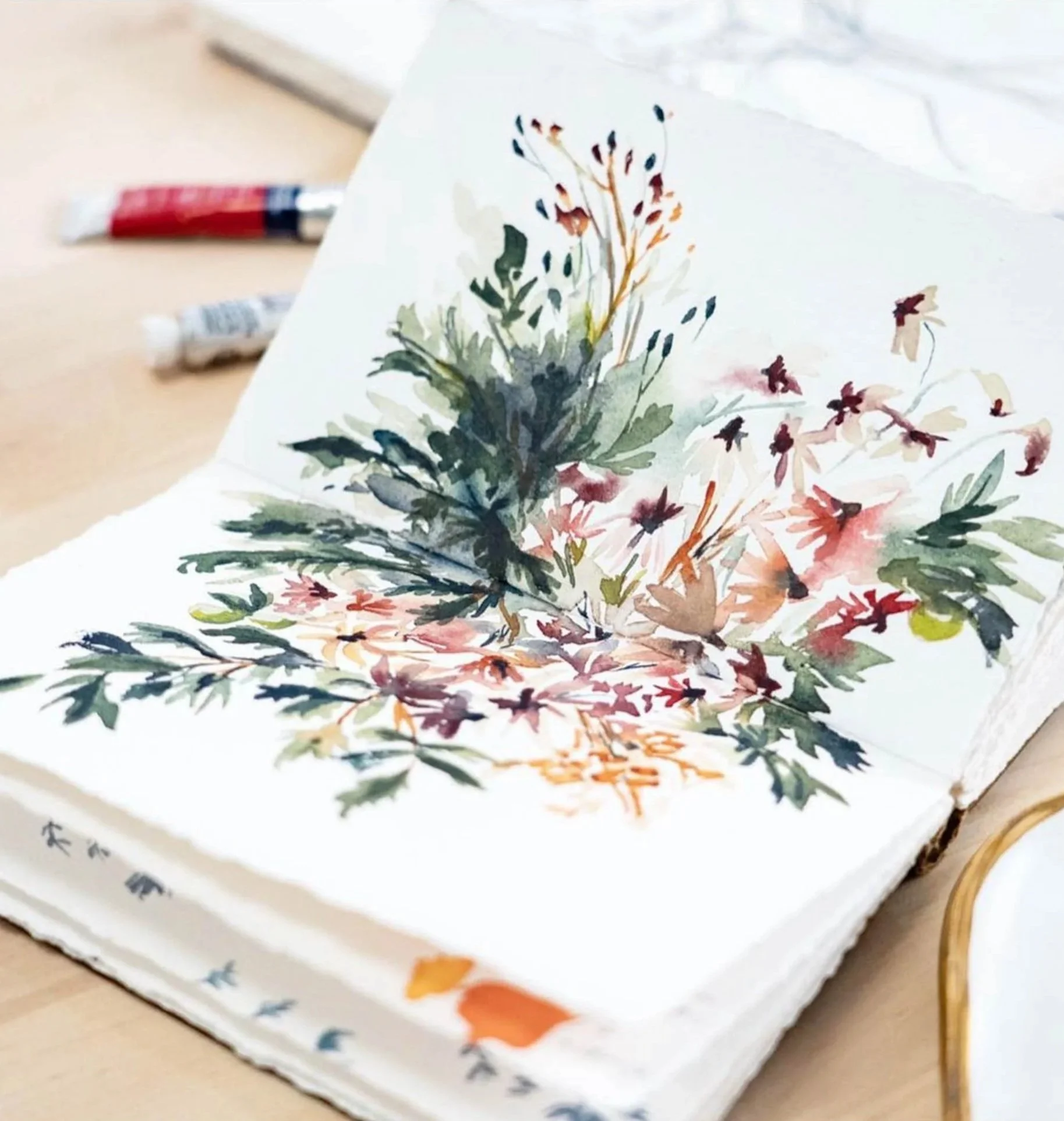 A watercolor painting of a bouquet of different plants and flowers on paper, with two tubes of red and white paint in the background, placed on a light-colored surface.