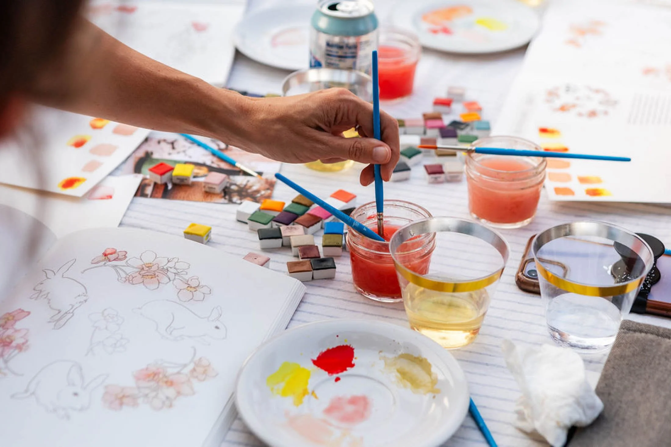 A person’s hand holding paintbrushes over a table filled with watercolor paints, paper with floral sketches, and art supplies.