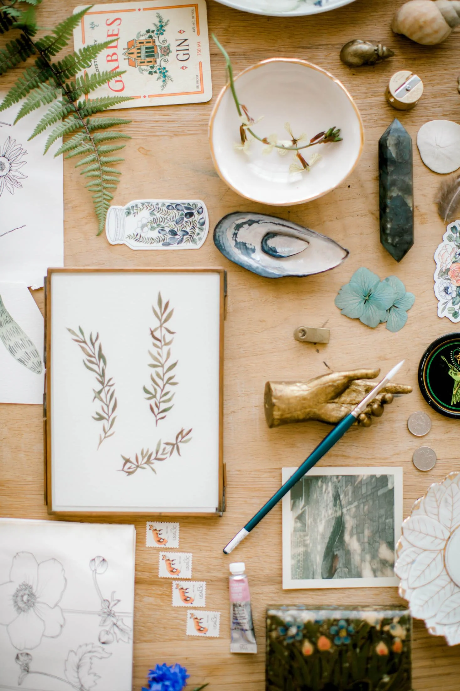 A flat lay of arts and crafts supplies on a wooden surface, including sketches of flowers, a watercolor painting, stamps, a tube of paint, seashells, a feather, a decorated stone, a small plant illustration, and a piece of driftwood, with some botanical stickers and a green marker.