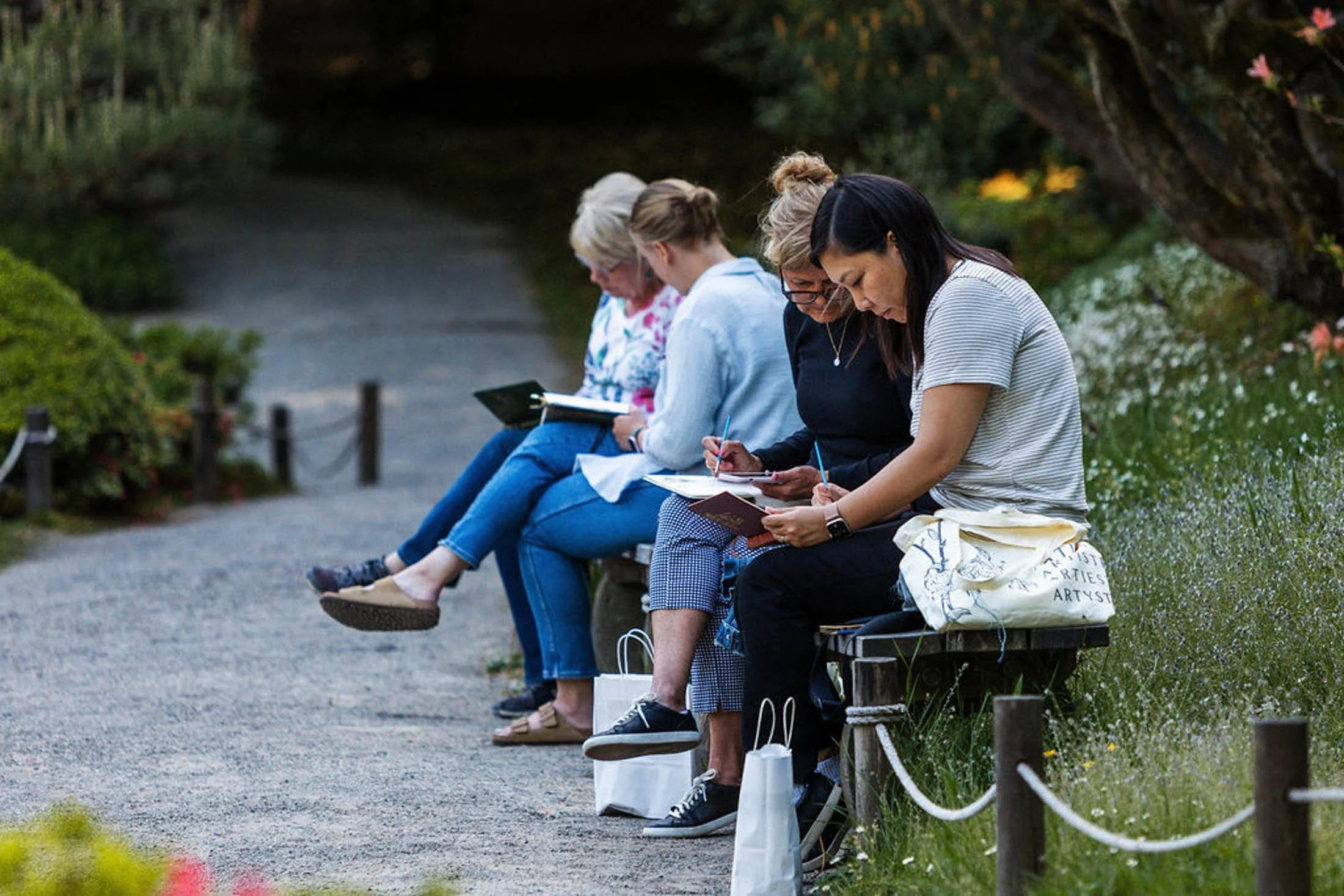 A group of five women sitting on a wooden bench outdoors, writing or drawing in notebooks, with a bridge and greenery in the background.