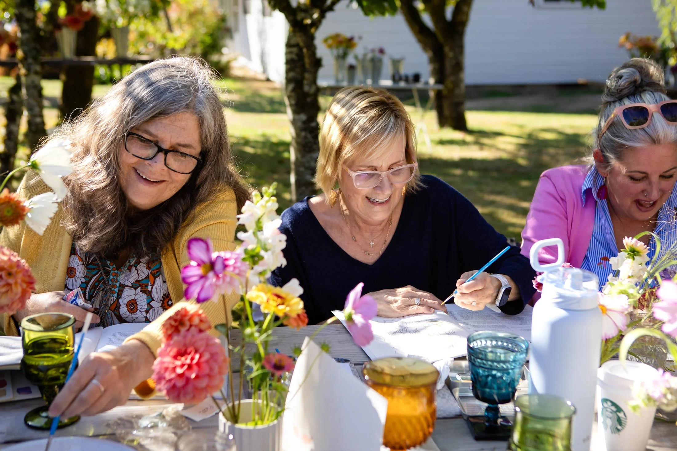 Three women smiling and sitting at a table outdoors, surrounded by colorful flowers, with trees and a house in the background.